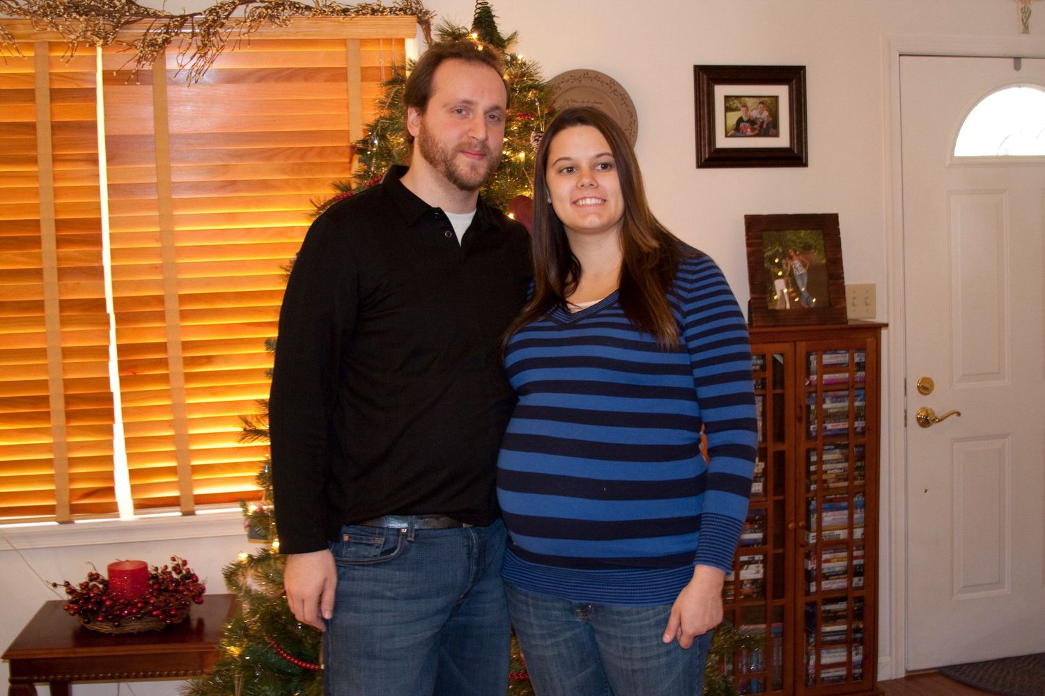 Couple in casual sweaters posing in front of window with holiday decorations.