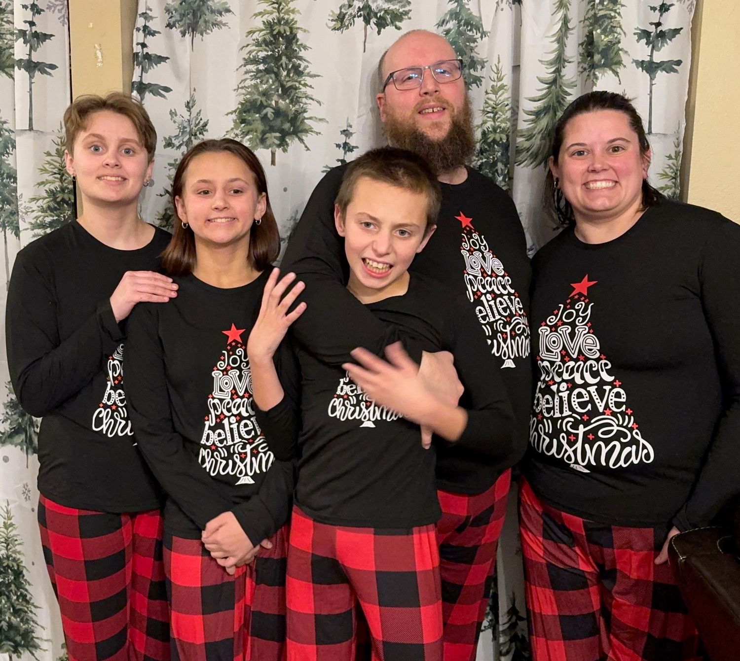 Group wearing matching black sweaters and buffalo plaid pajama pants against tree-patterned backdrop.