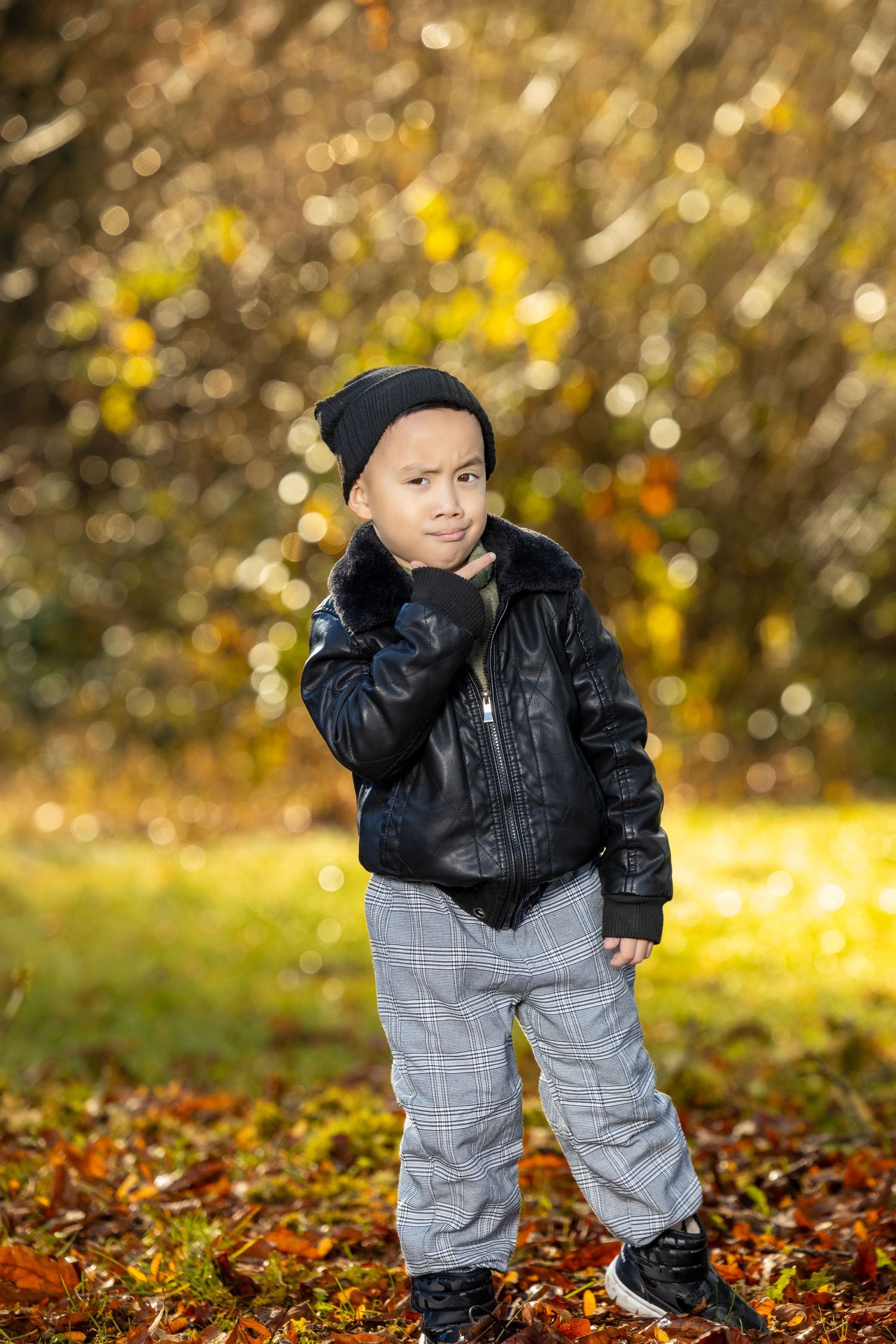 A child in a black jacket and gray pants plays in fallen autumn leaves with golden light in background.
