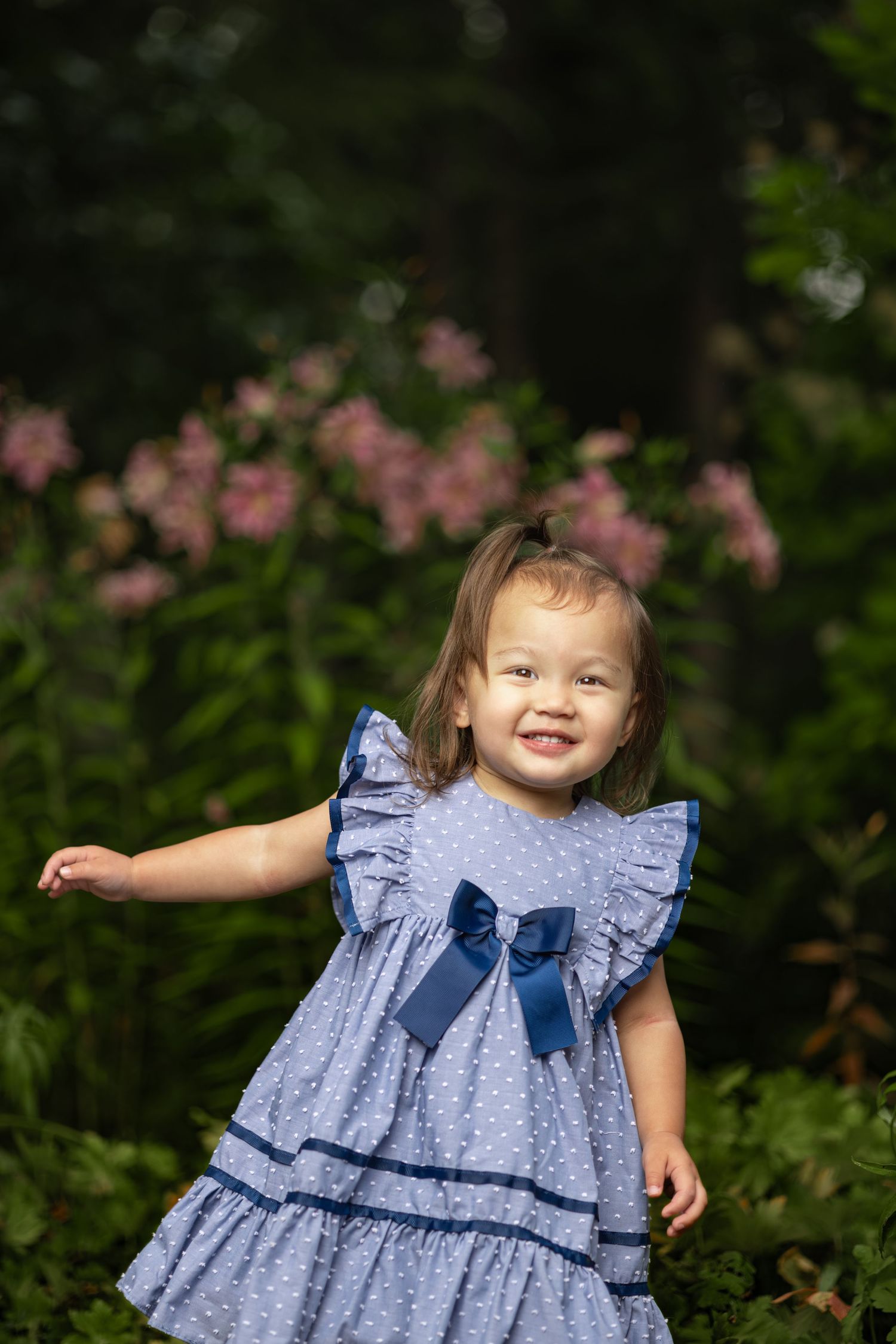 A child in a blue ruffled dress with a bow plays outdoors in a garden setting with pink flowers in the background.