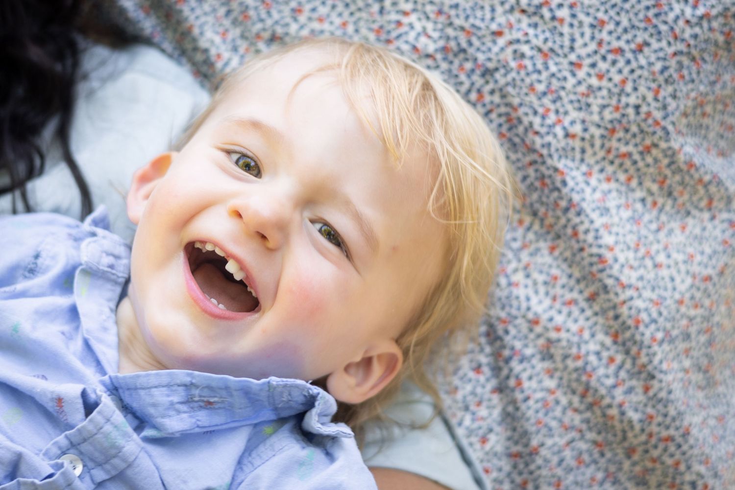 A joyful toddler with blonde hair laughs while lying on patterned bedding.