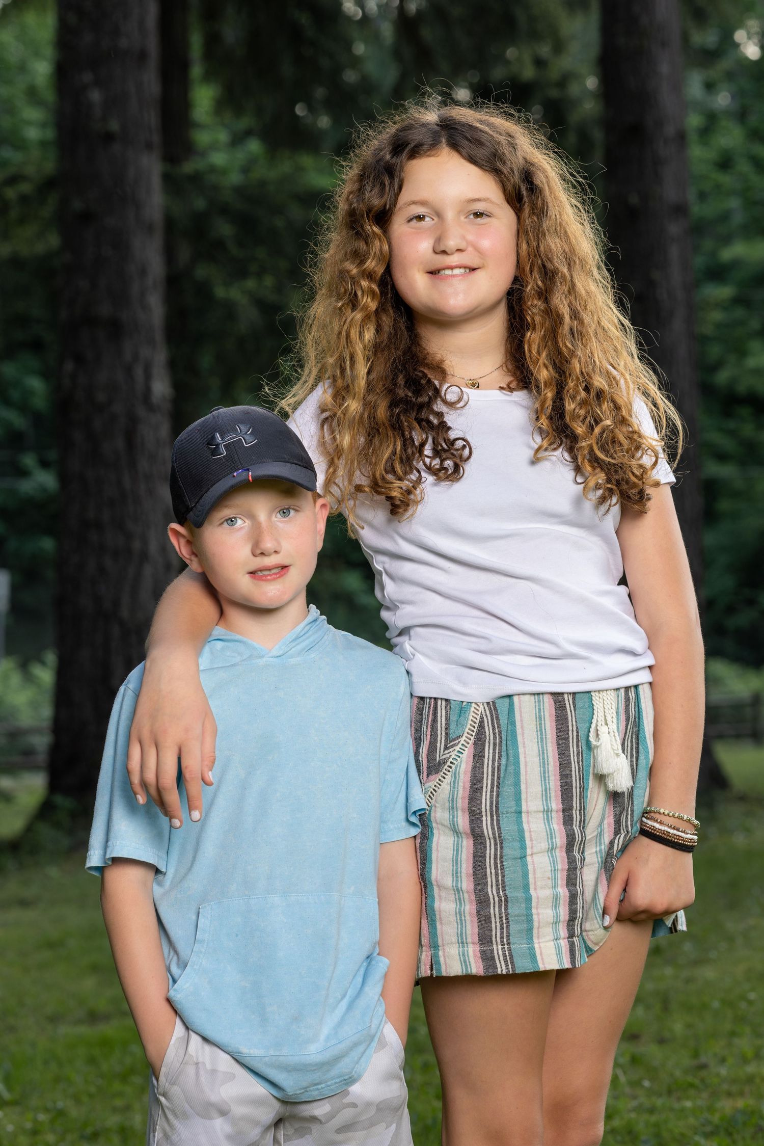 Children pose together outdoors wearing casual summer clothes among pine trees.