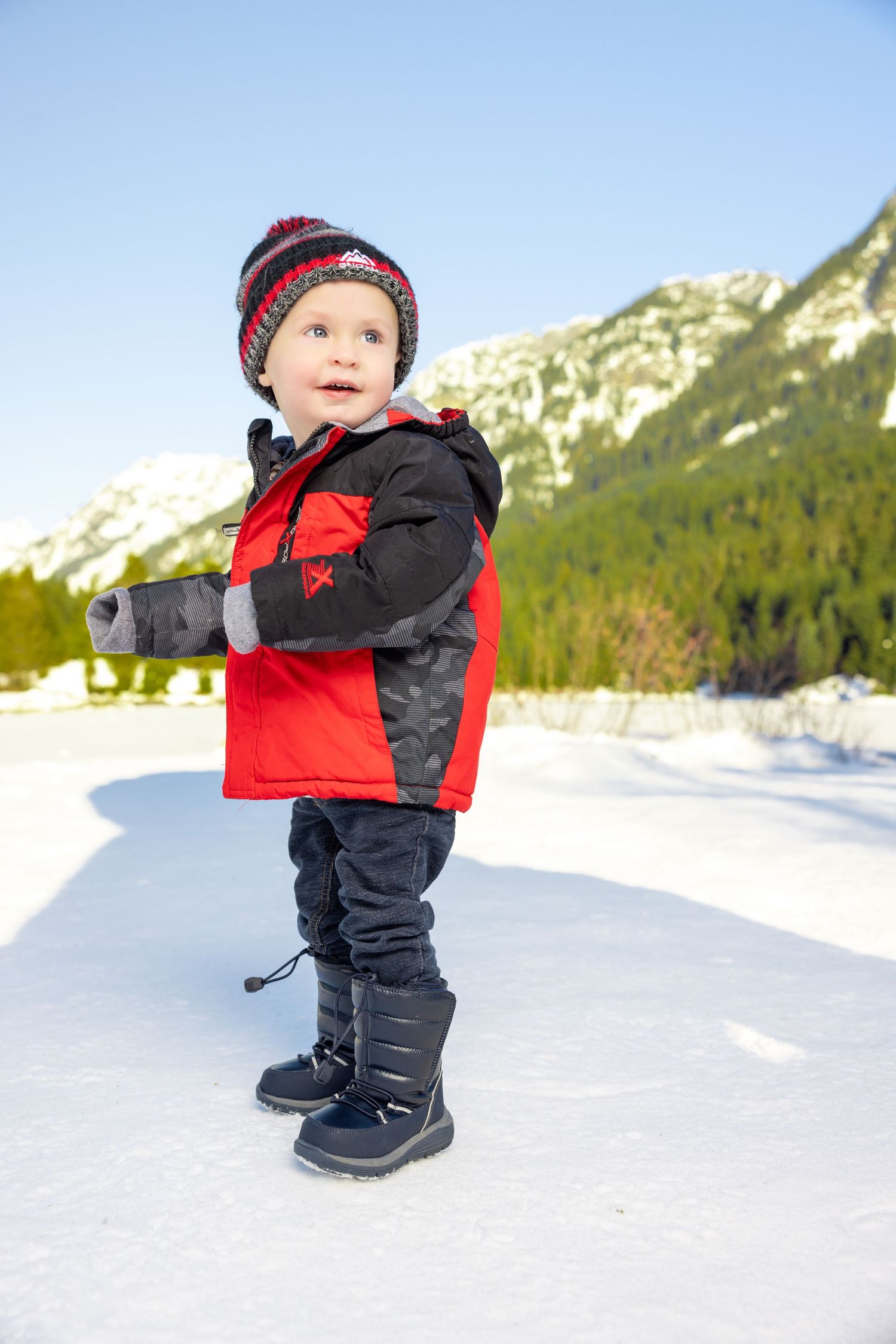 Small child in winter gear walking through deep snow with mountains in background.