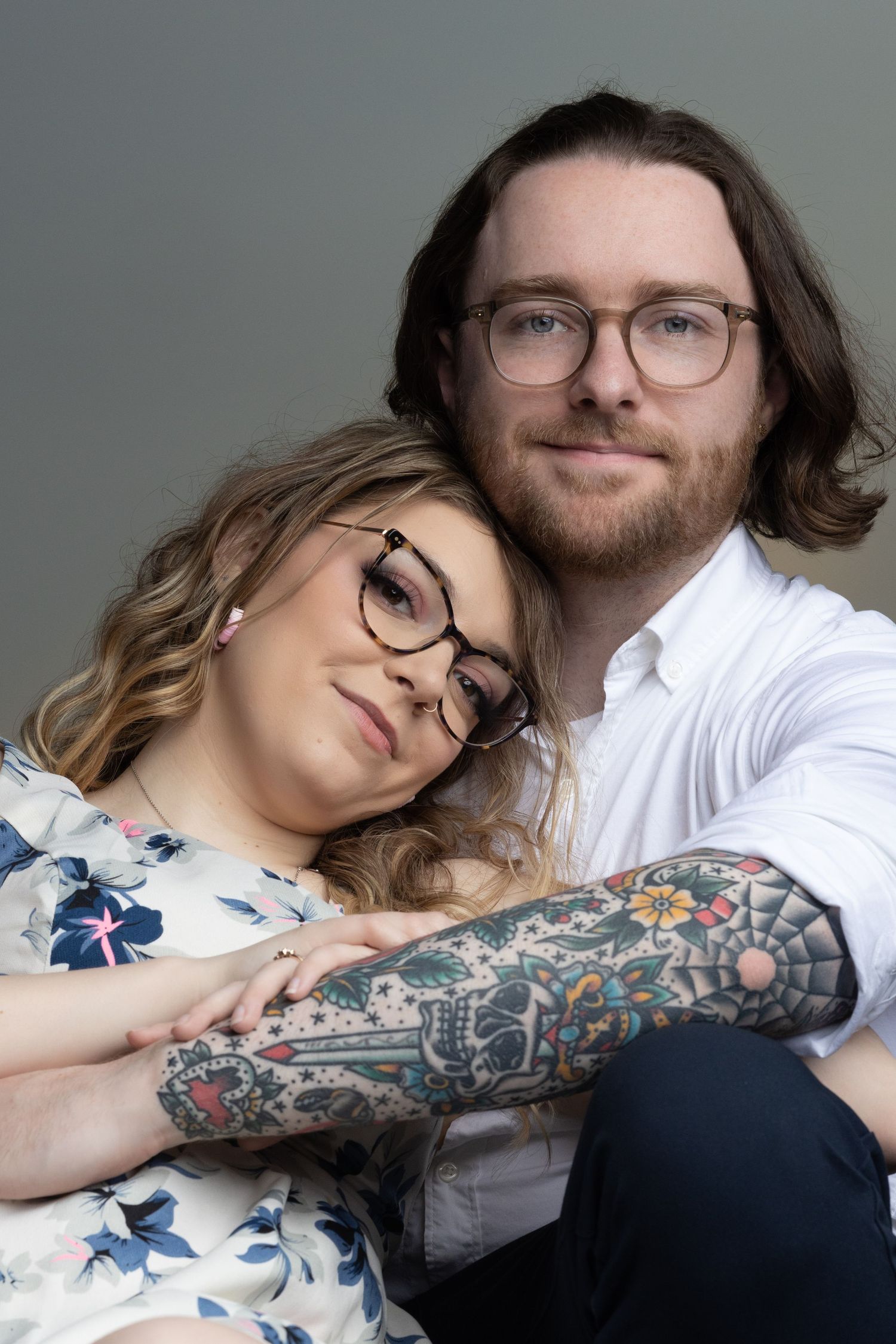 A couple in glasses embrace while showing off intricate floral sleeve tattoo artwork.