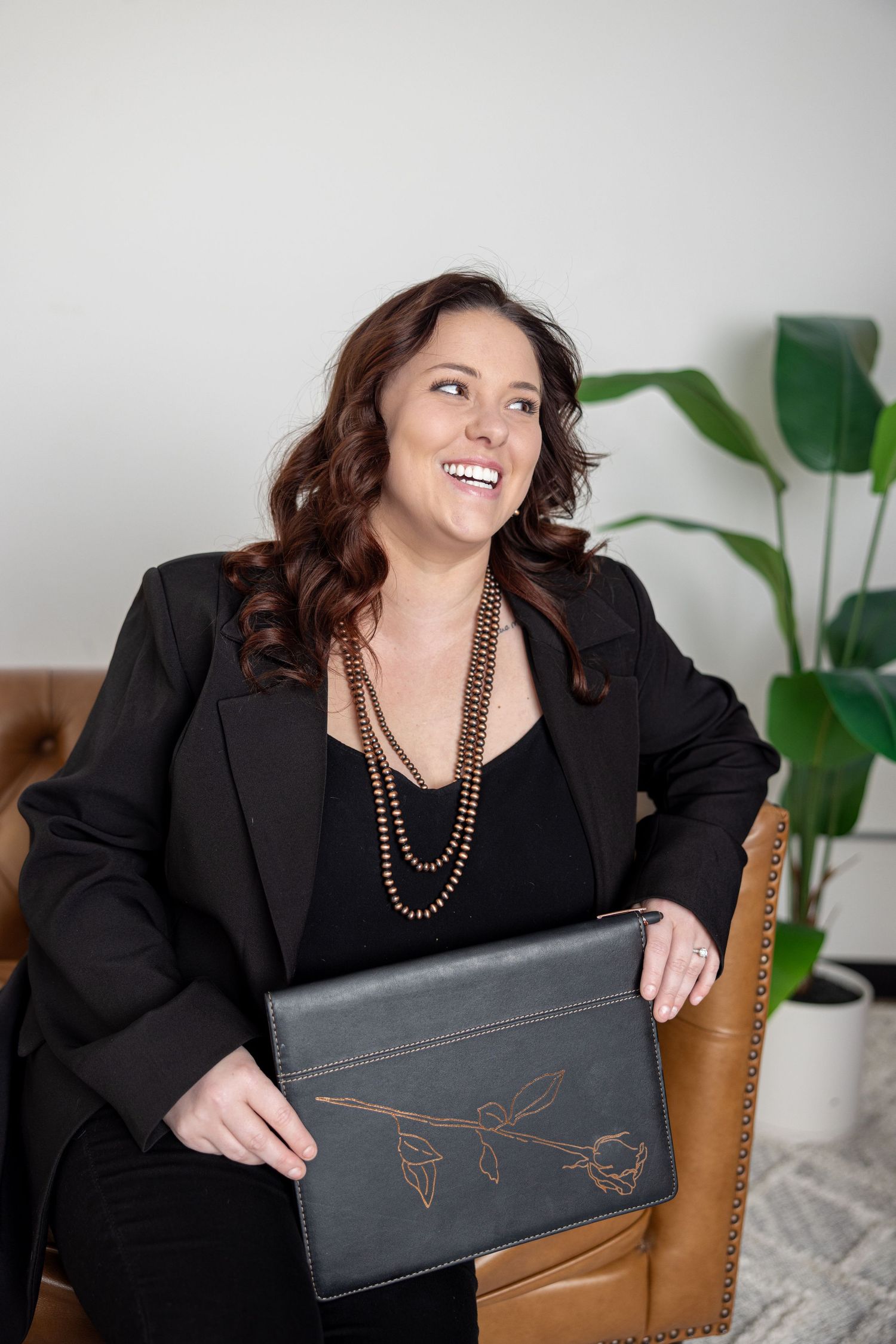 Woman in business attire poses with black portfolio on leather sofa.