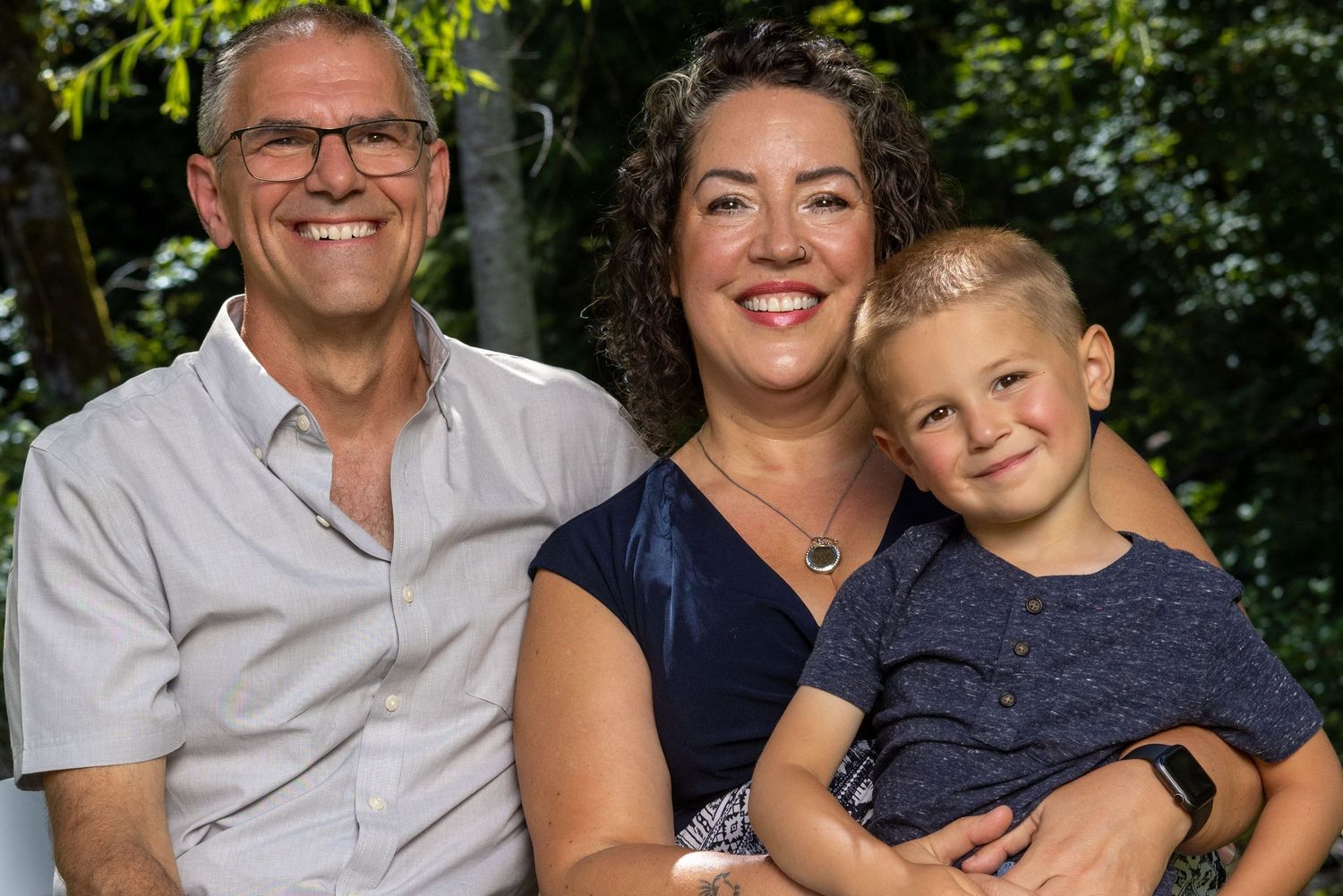 Happy family of three poses together outdoors on a sunny day.
