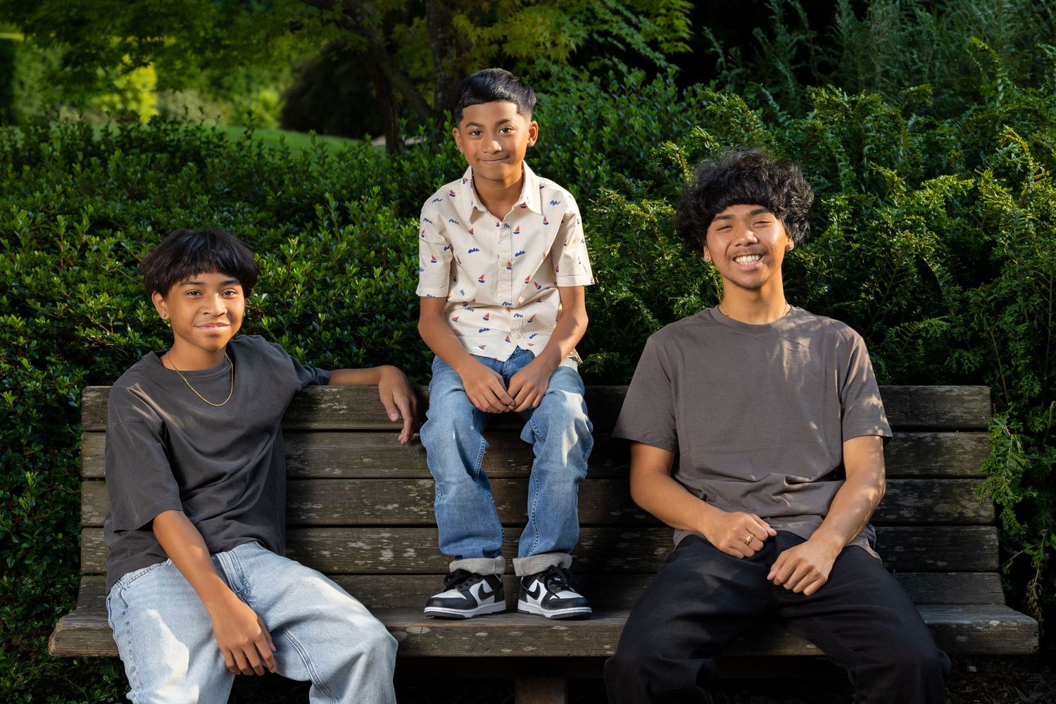 Three youth sitting casually on wooden bench in garden setting.