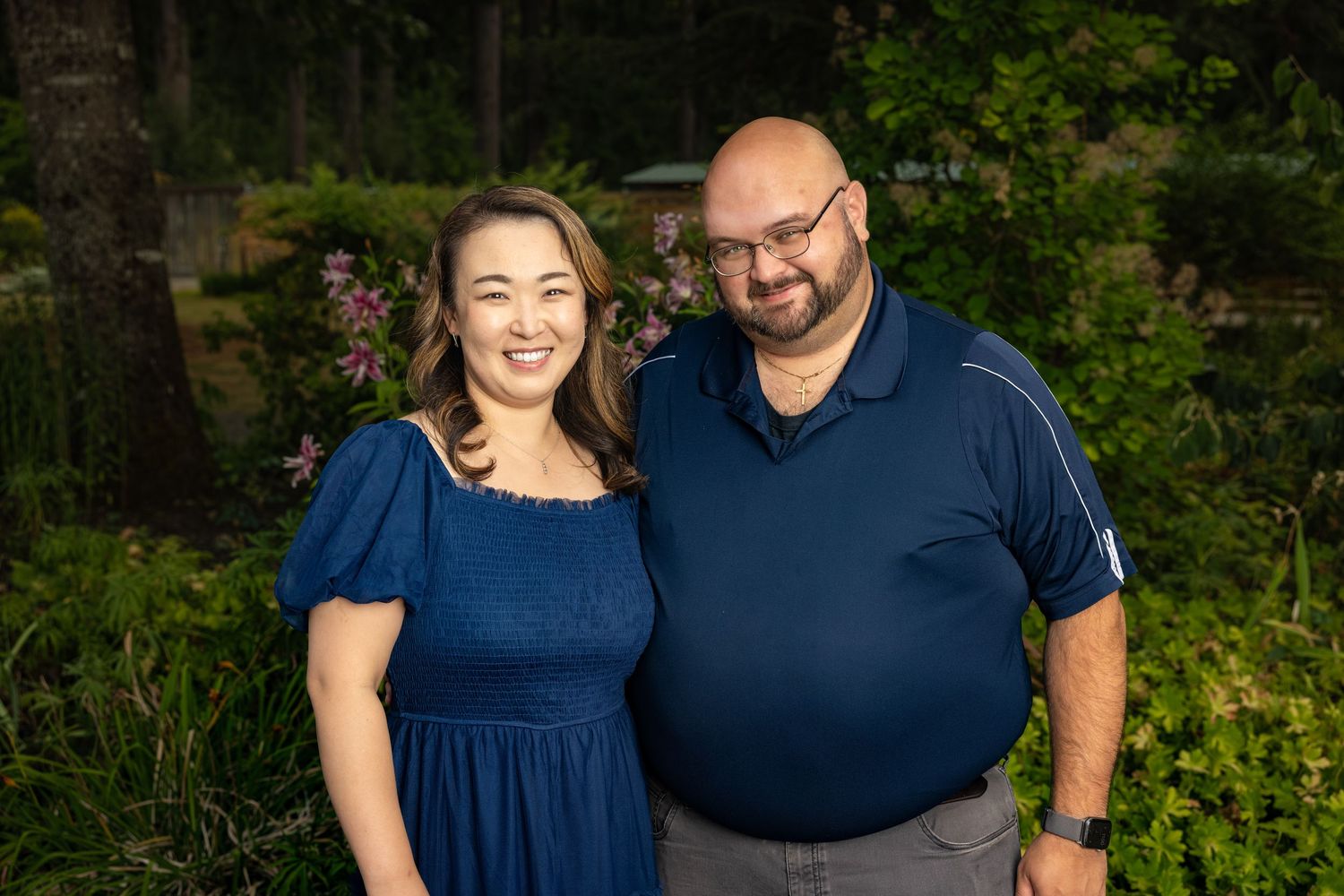 A couple wearing coordinating navy blue outfits poses together outdoors in a garden setting.