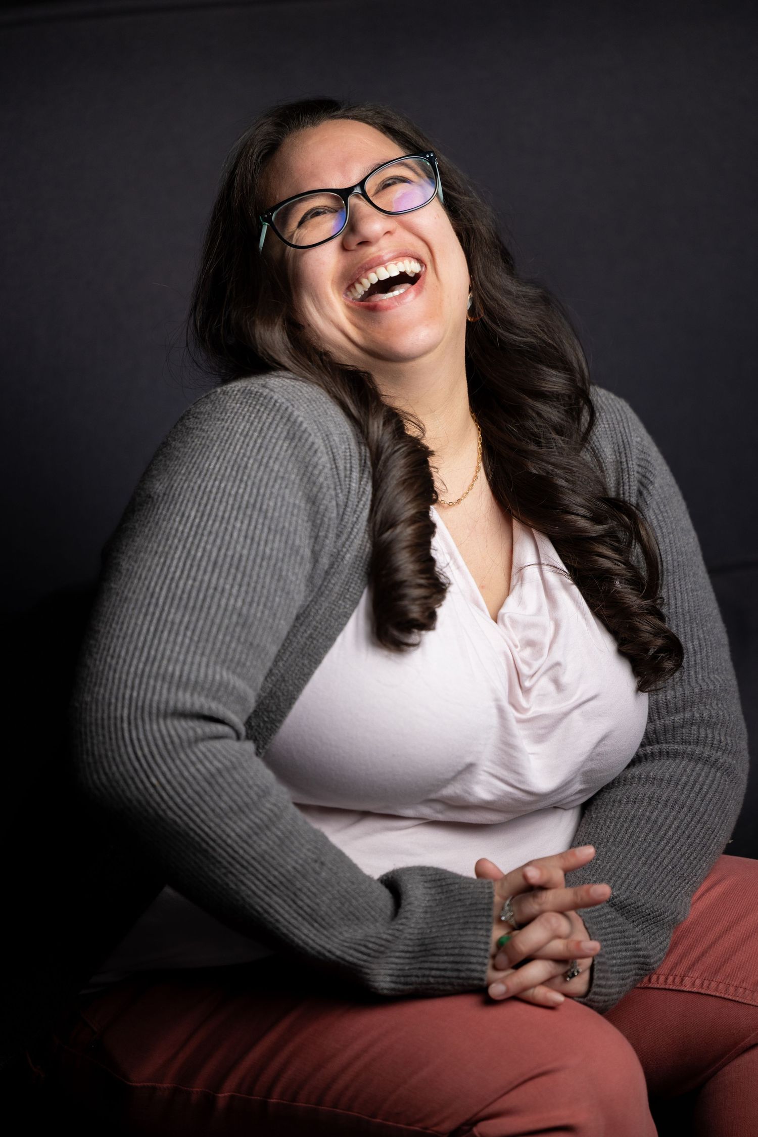 Portrait of someone in glasses and gray sweater laughing joyfully against dark background.