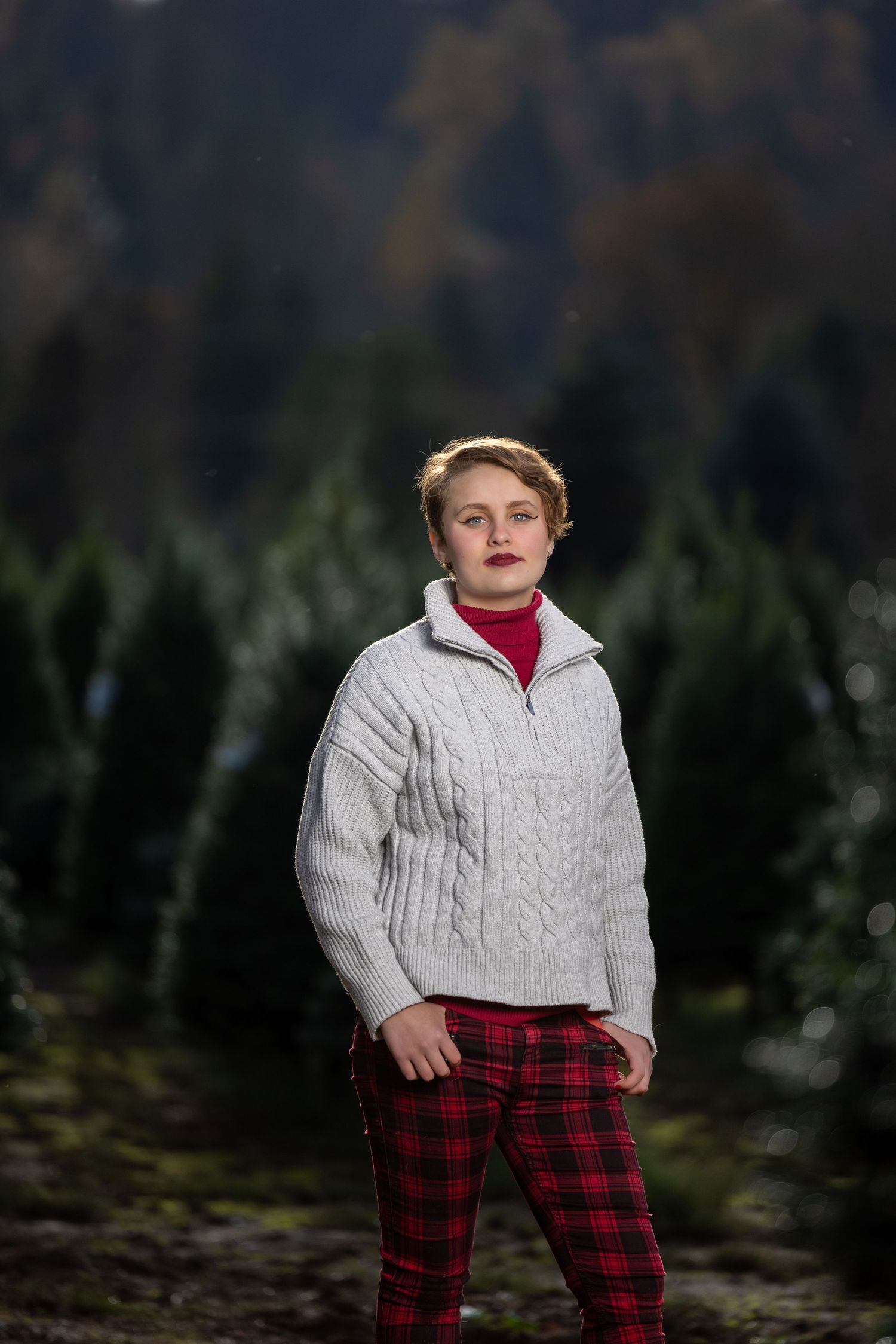 Person wearing plaid pants and cable knit sweater stands in front of forest backdrop.