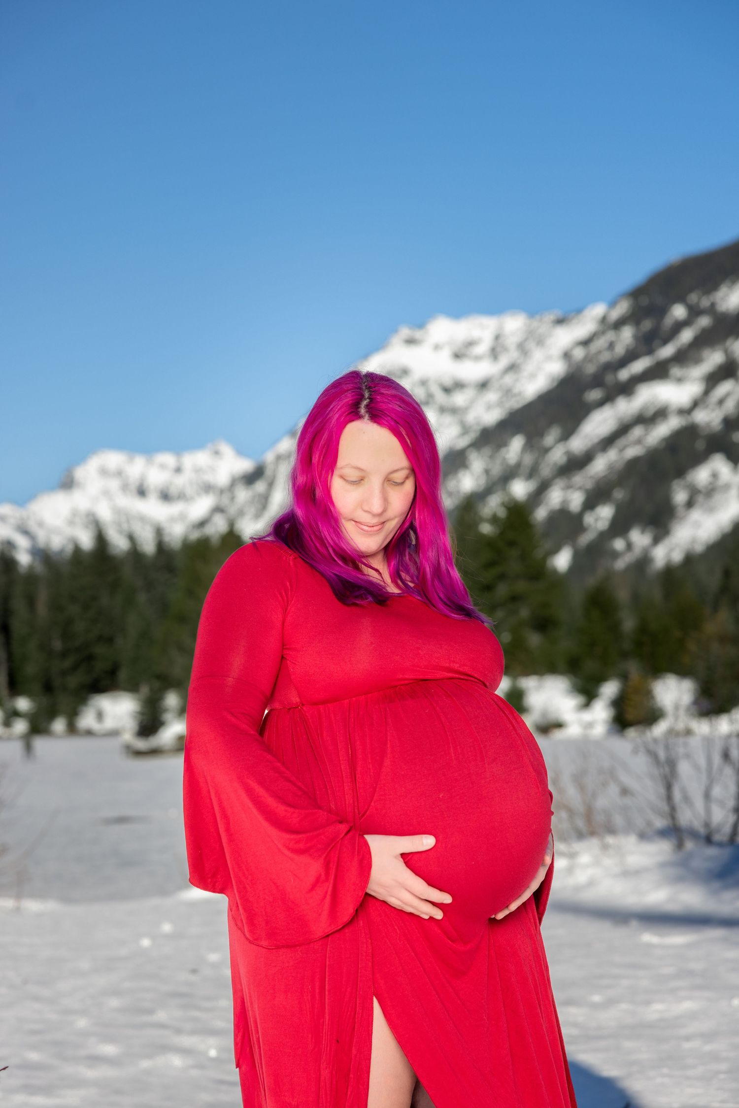Maternity photos in red dress against snowy mountain landscape.