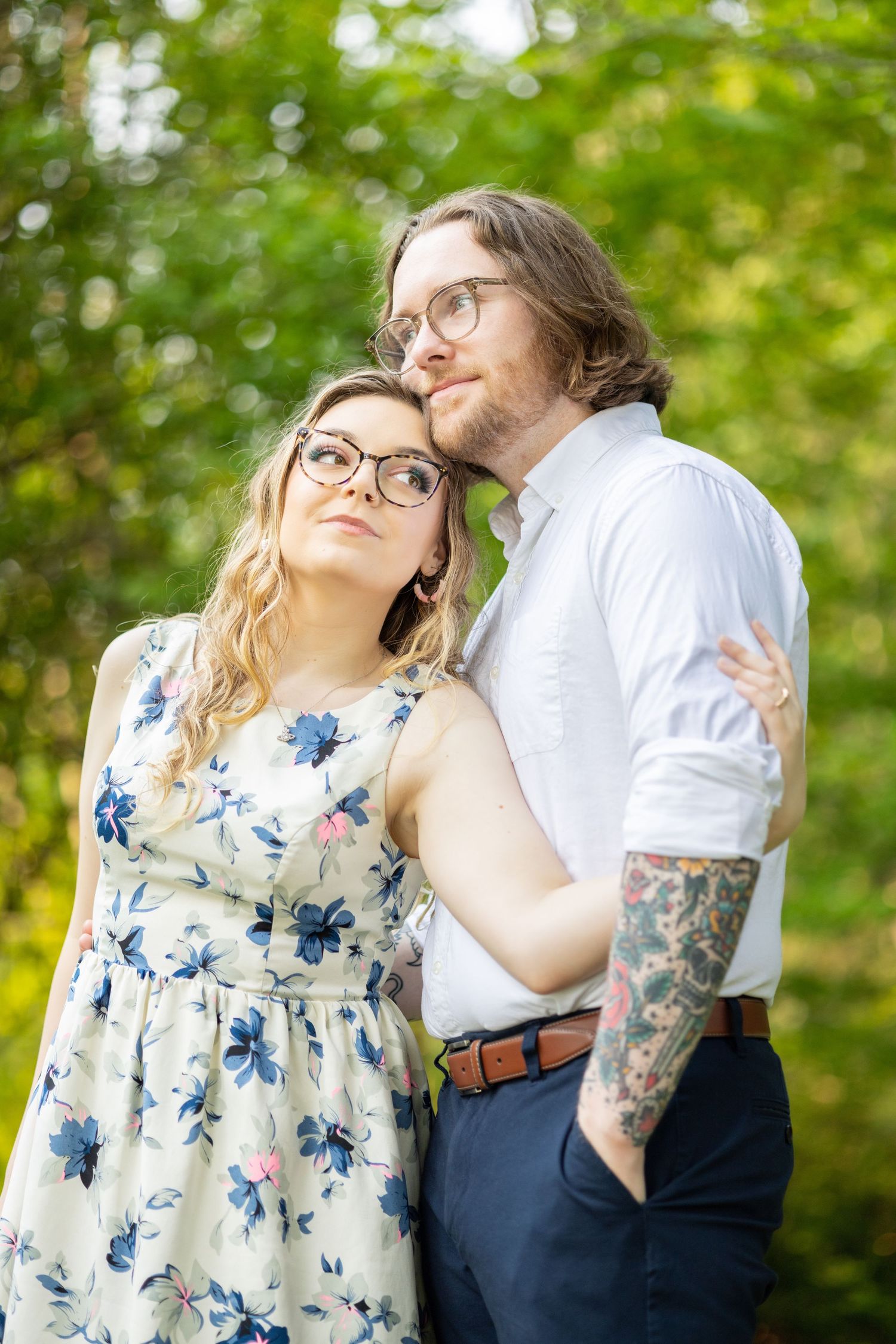 Romantic outdoor couple portrait with floral dress and visible tattoo sleeve in sunlit garden setting.