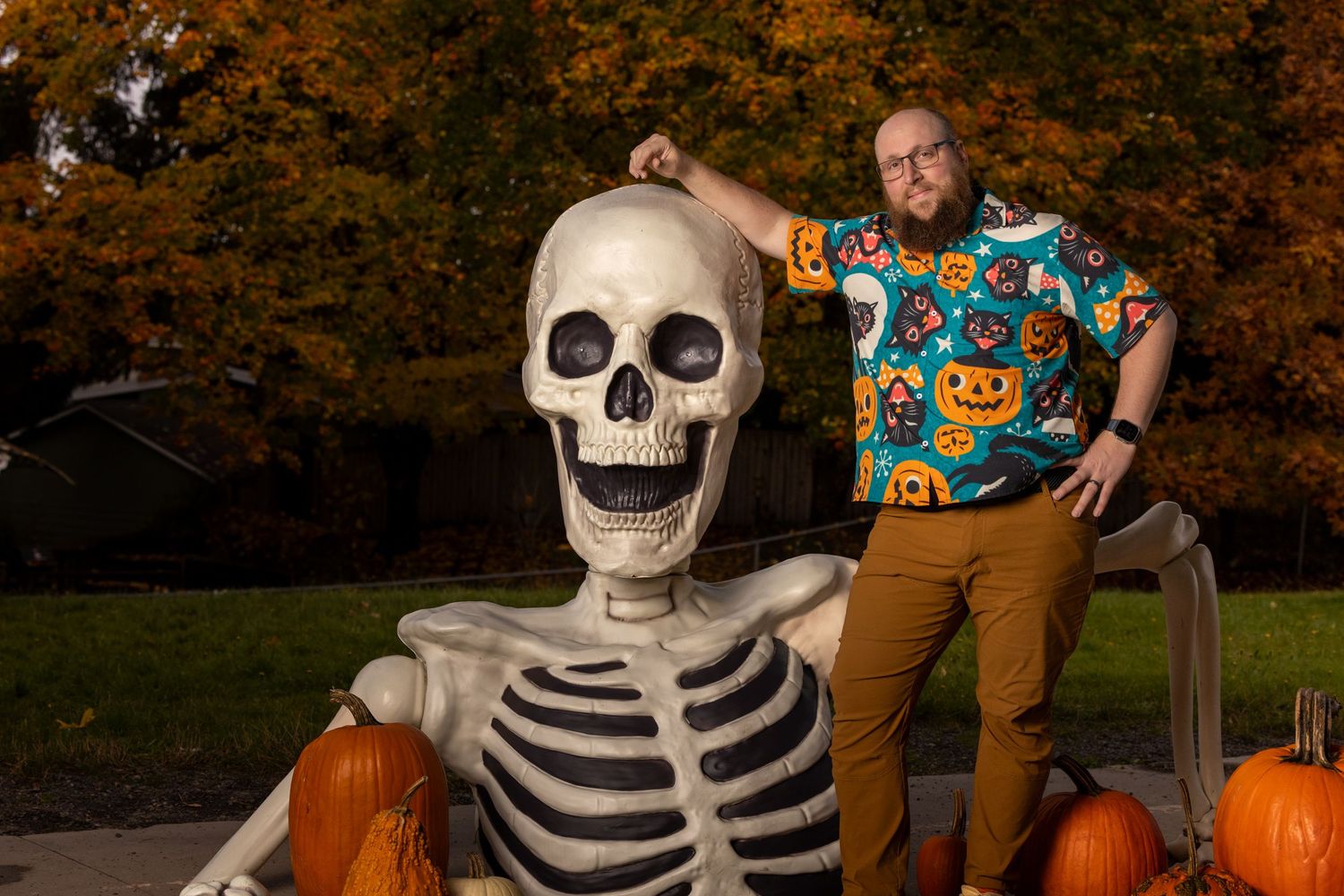 A person in a Halloween-themed shirt poses next to a large decorative skeleton and pumpkins in an autumn outdoor setting.