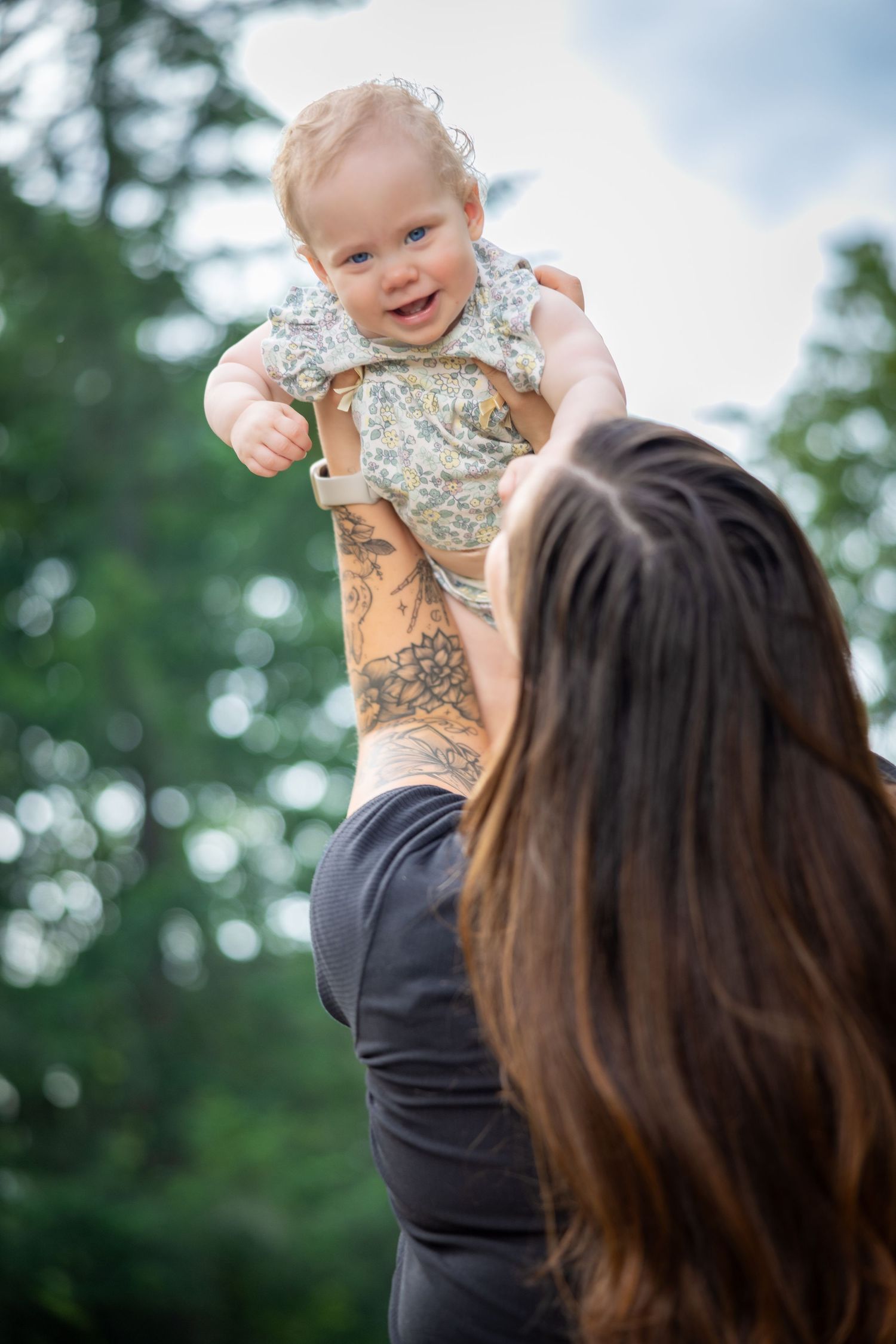 Baby in floral outfit being lifted high in outdoor setting with trees in background.