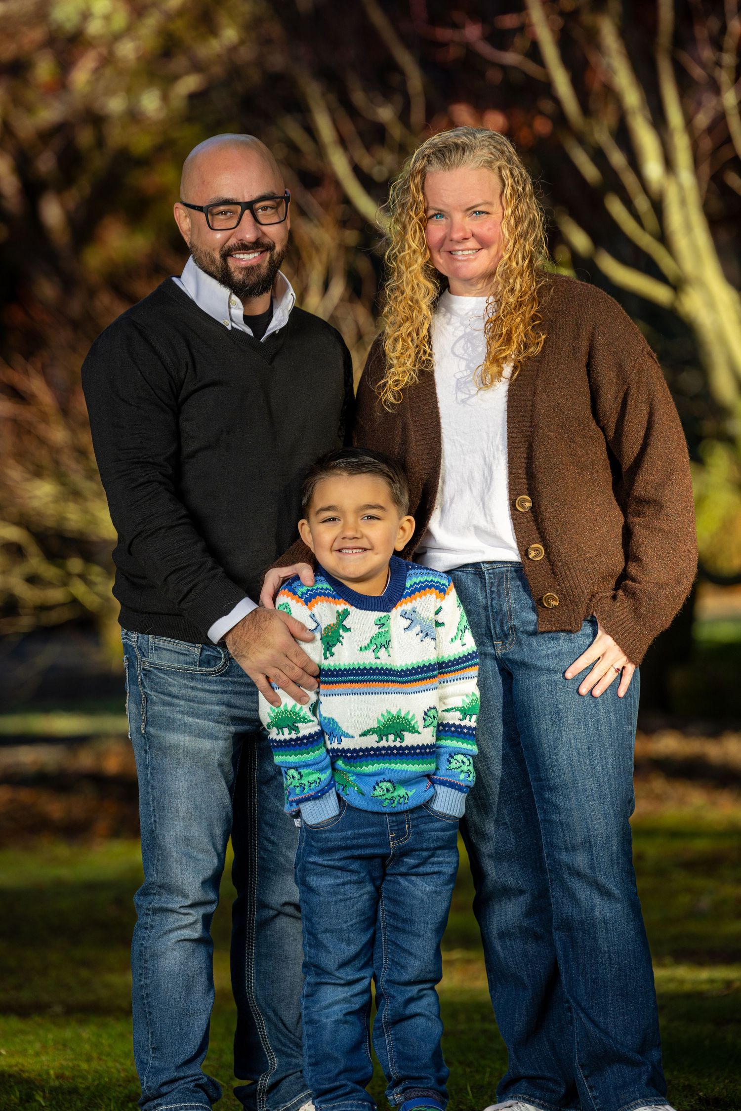 Family portrait in casual attire featuring denim and a dinosaur-themed sweater outdoors.