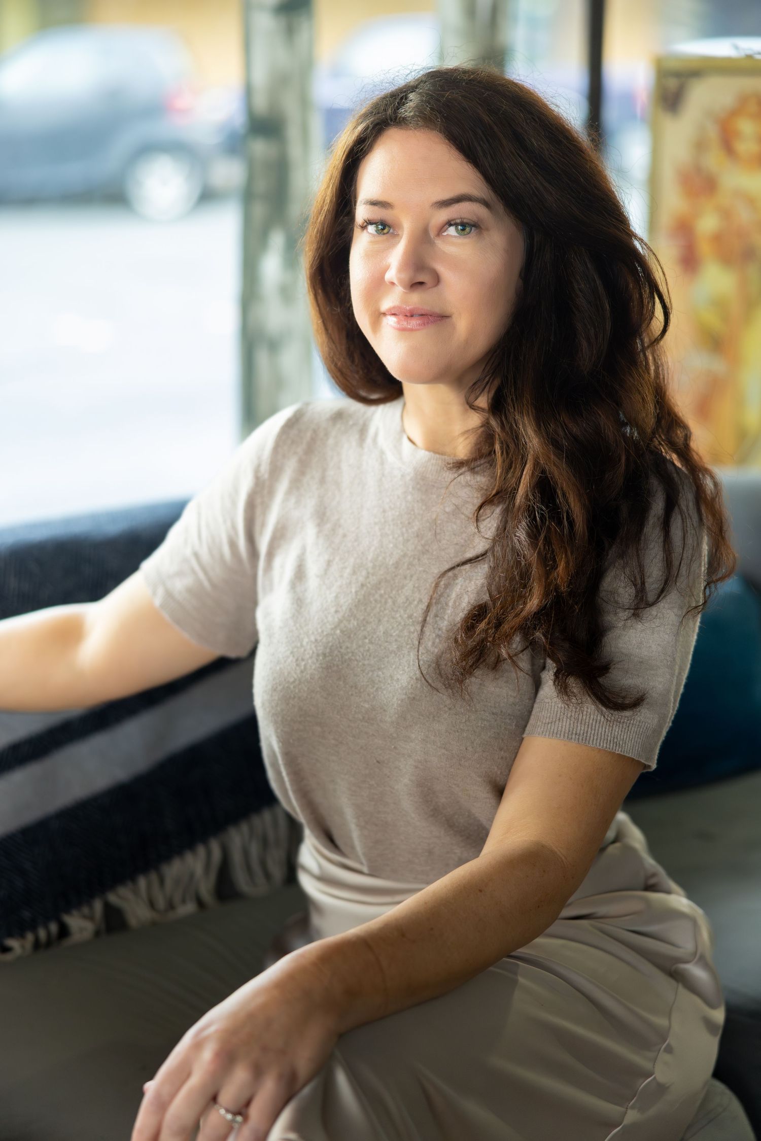 Woman in beige top sitting casually by window in natural lighting.