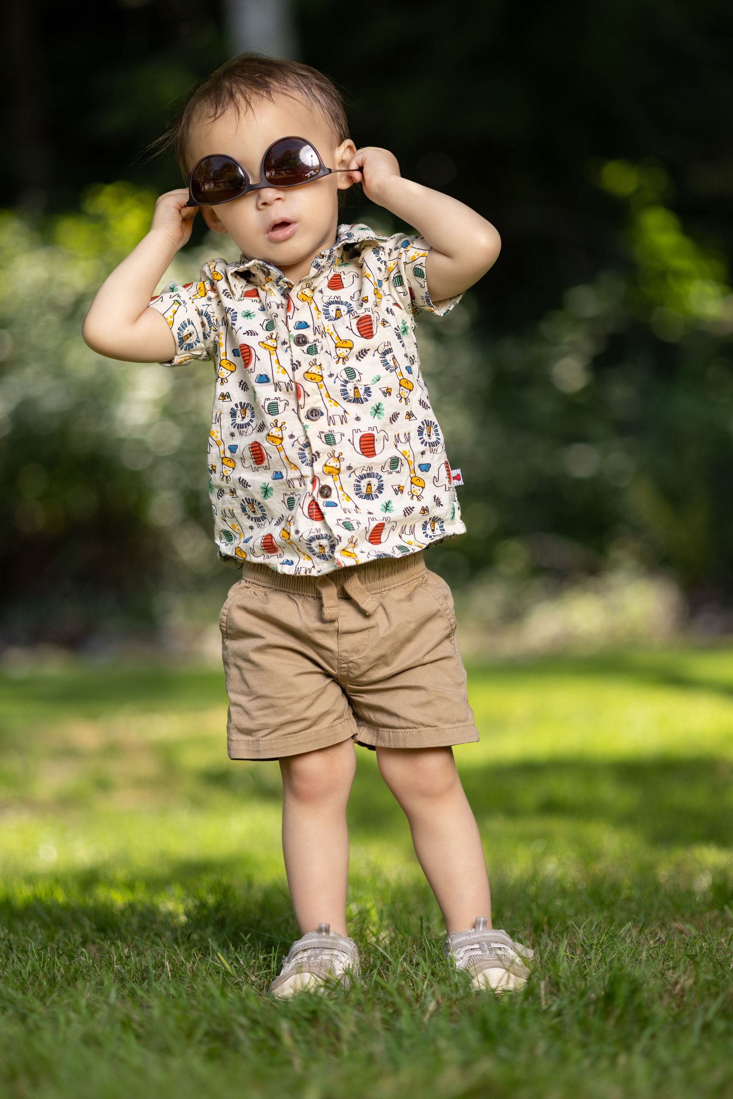 Small child in patterned shirt and khaki shorts wearing oversized sunglasses outdoors.