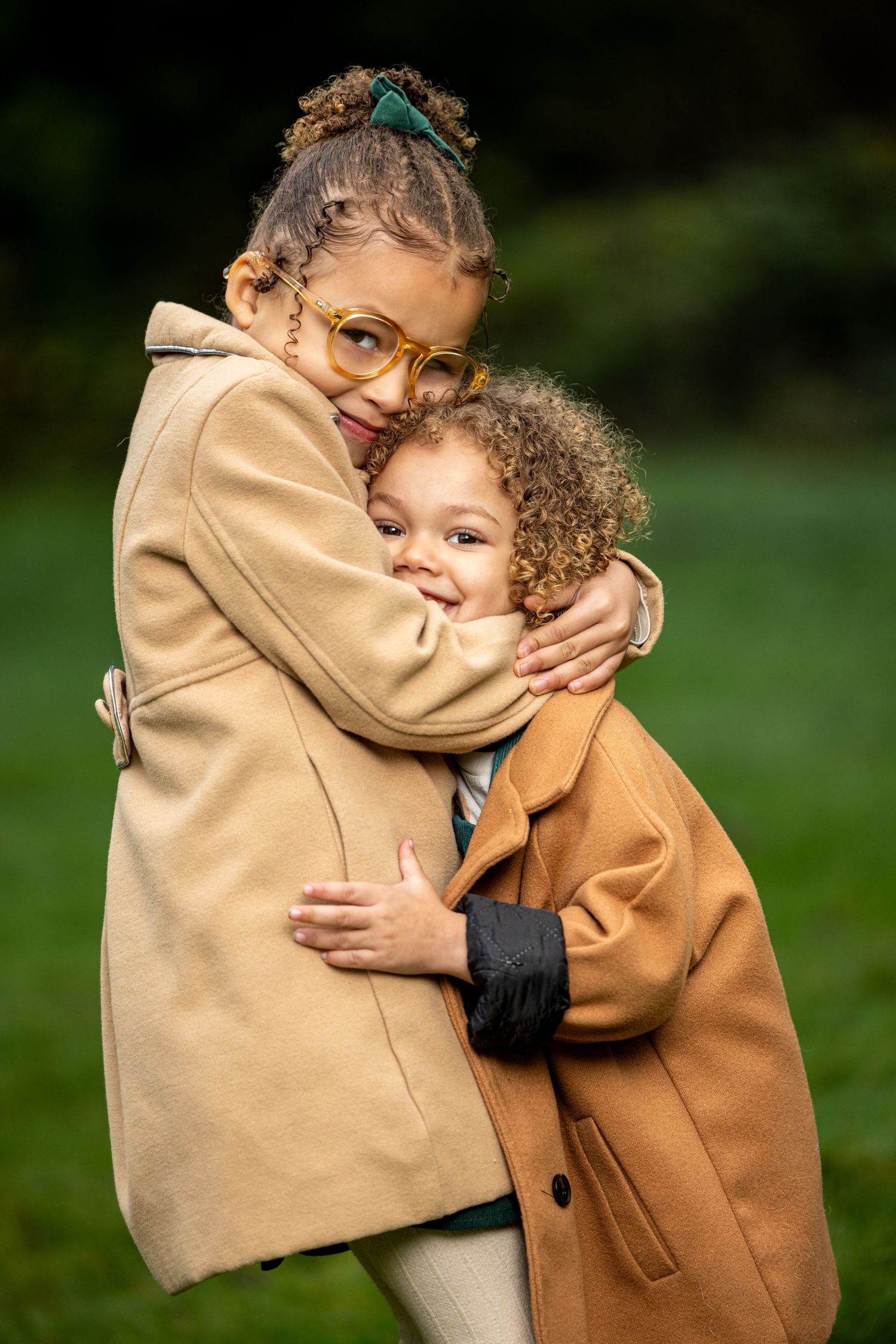 Two children in matching tan coats sharing a warm embrace outdoors.