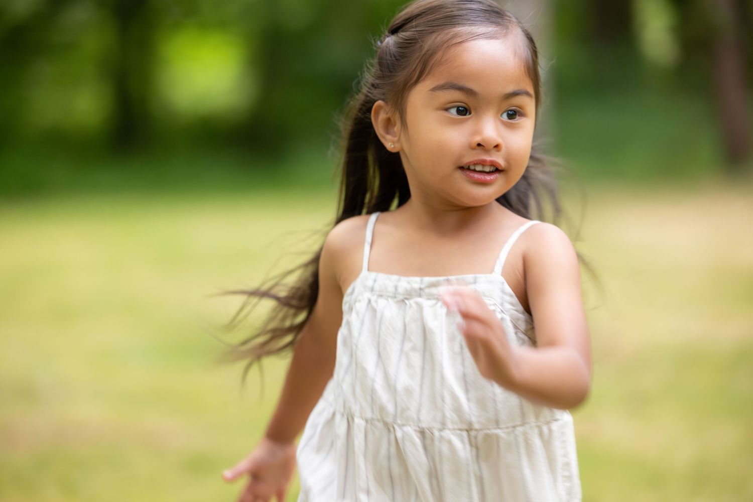 A child in a white summer dress runs and plays in an outdoor setting with trees in the background.
