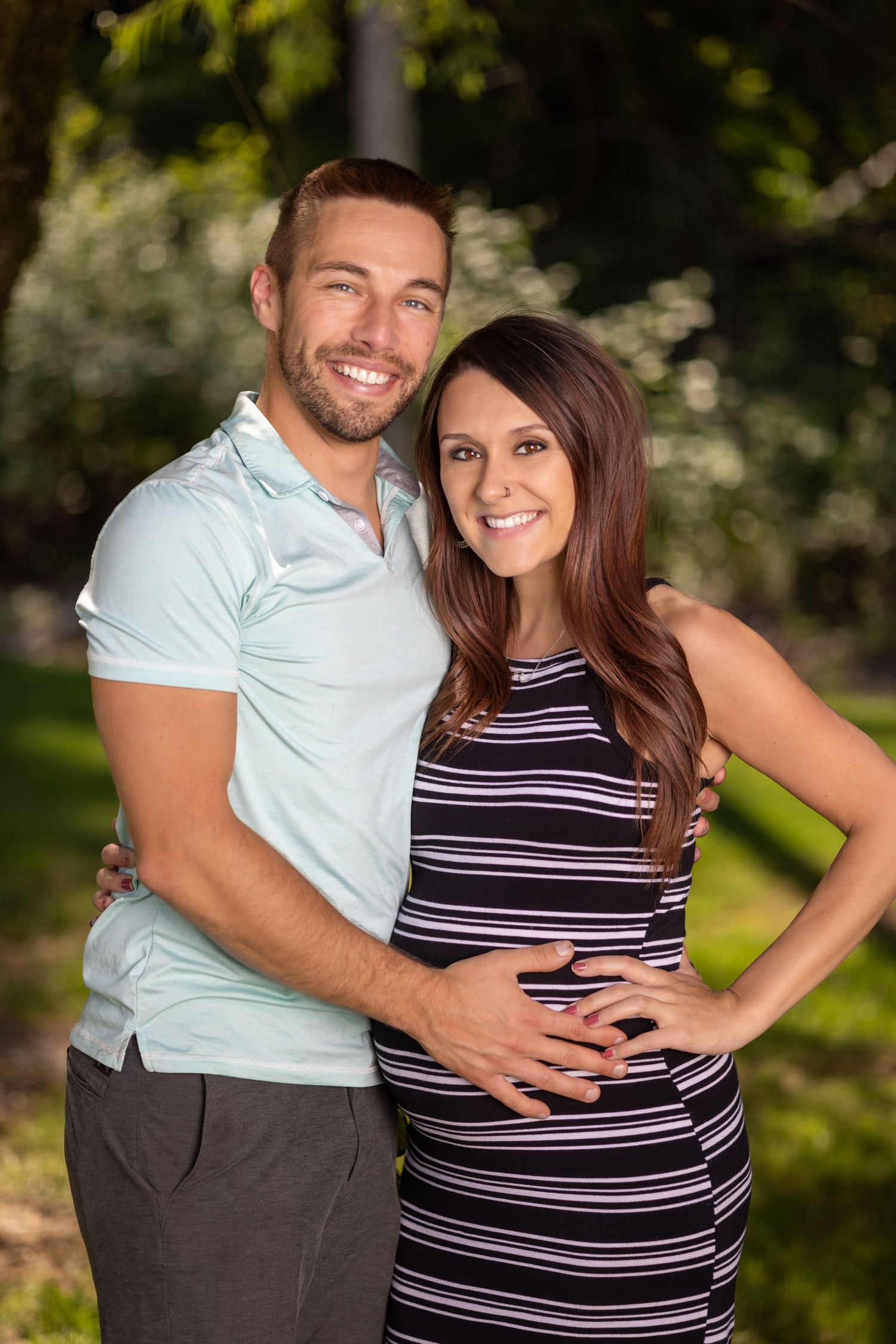 Couple embraces outdoors, with woman wearing striped dress showing baby bump.