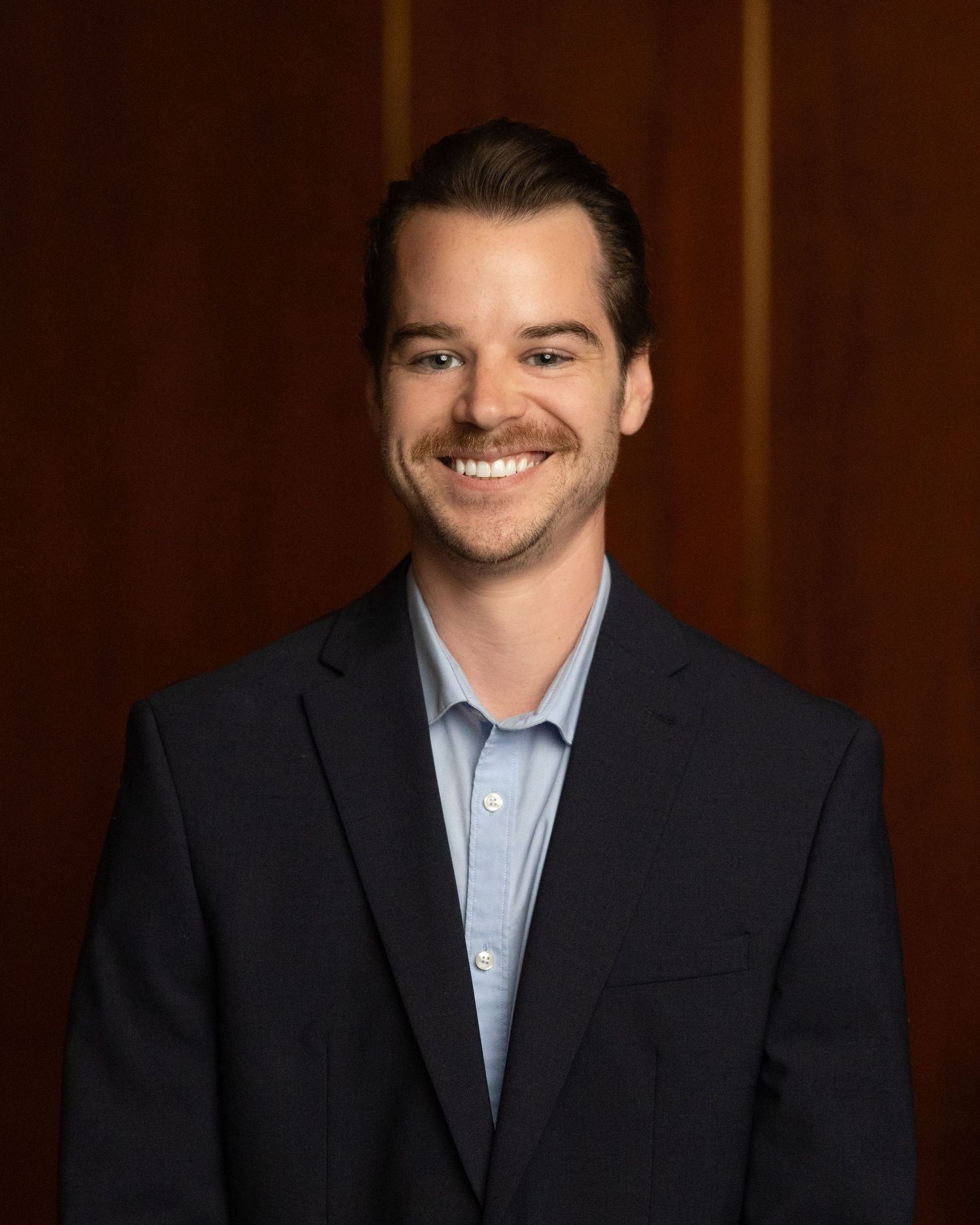 Professional headshots of person in dark suit and blue shirt against wood paneling.