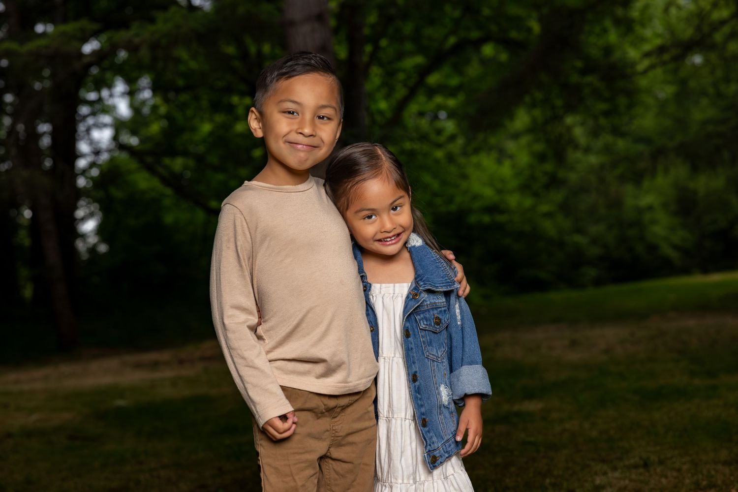 Two children embrace while standing outdoors in casual clothing against a natural background.