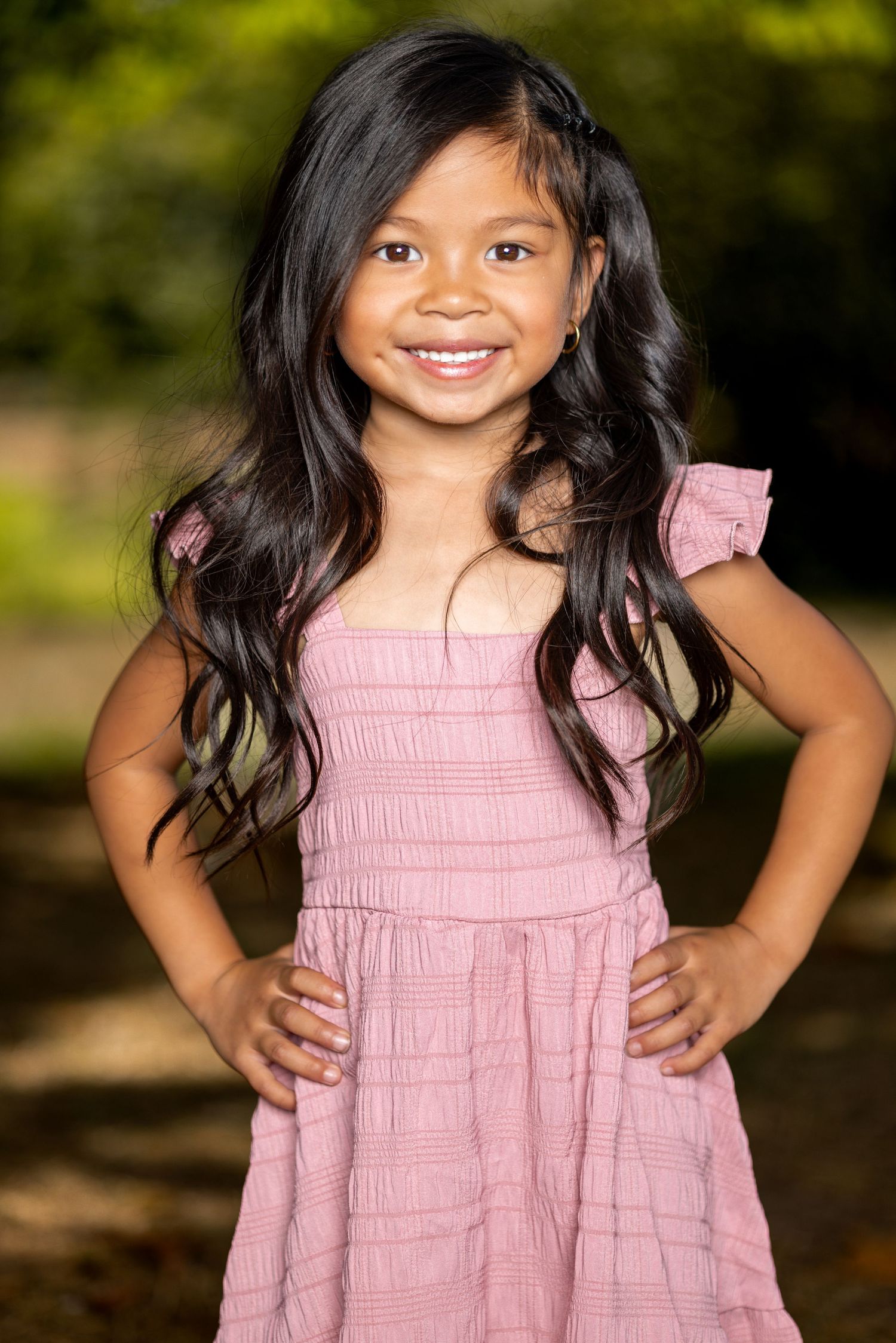 A child in a pink smocked dress poses outdoors with natural lighting and greenery in background.