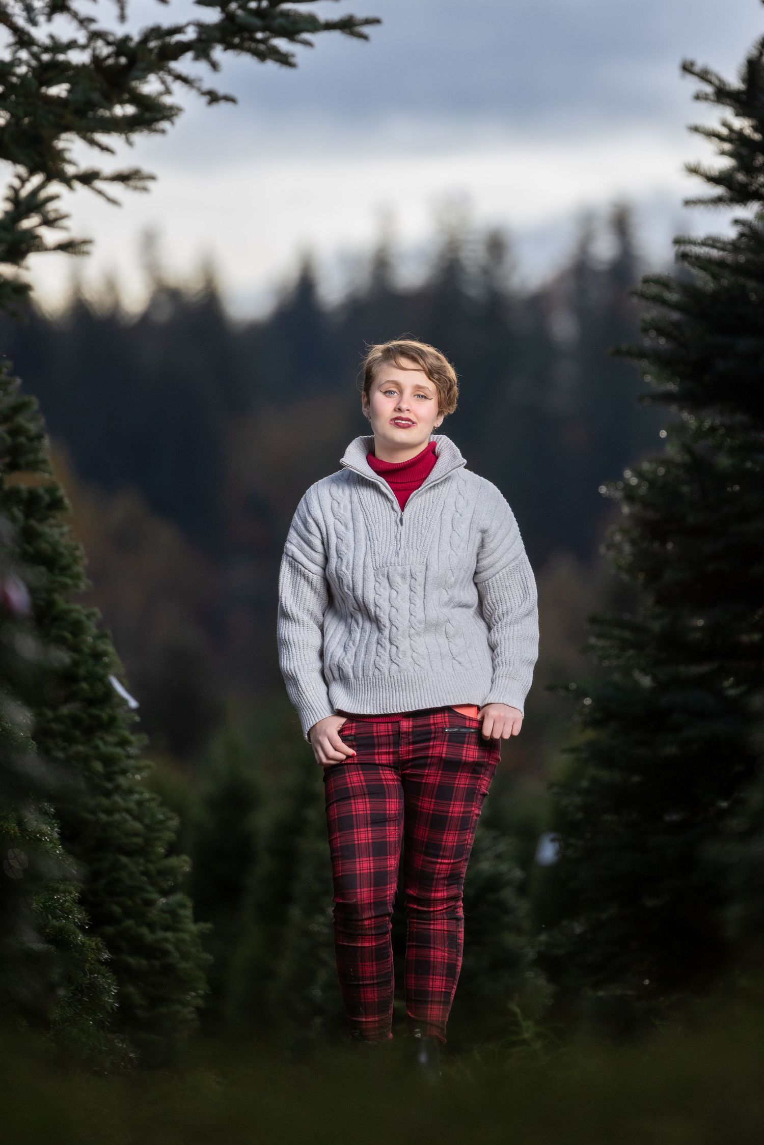 Person in red plaid pants and white sweater poses outdoors among evergreen trees.