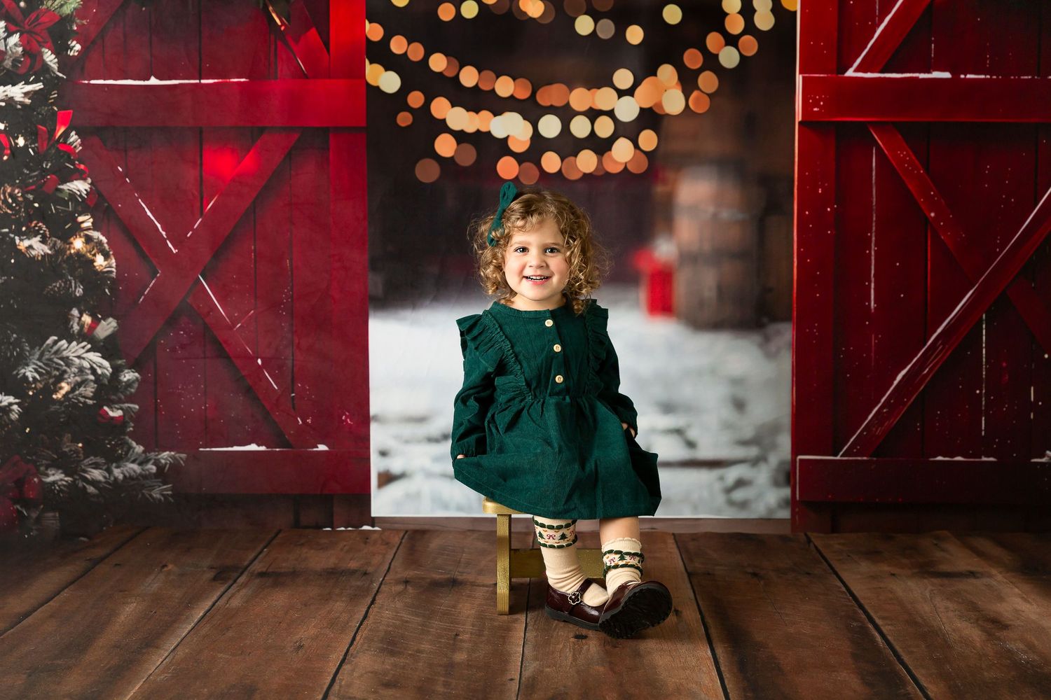 Toddler girl in green dress poses in front of festive red barn doors with bokeh lights and snowy Christmas tree backdrop.