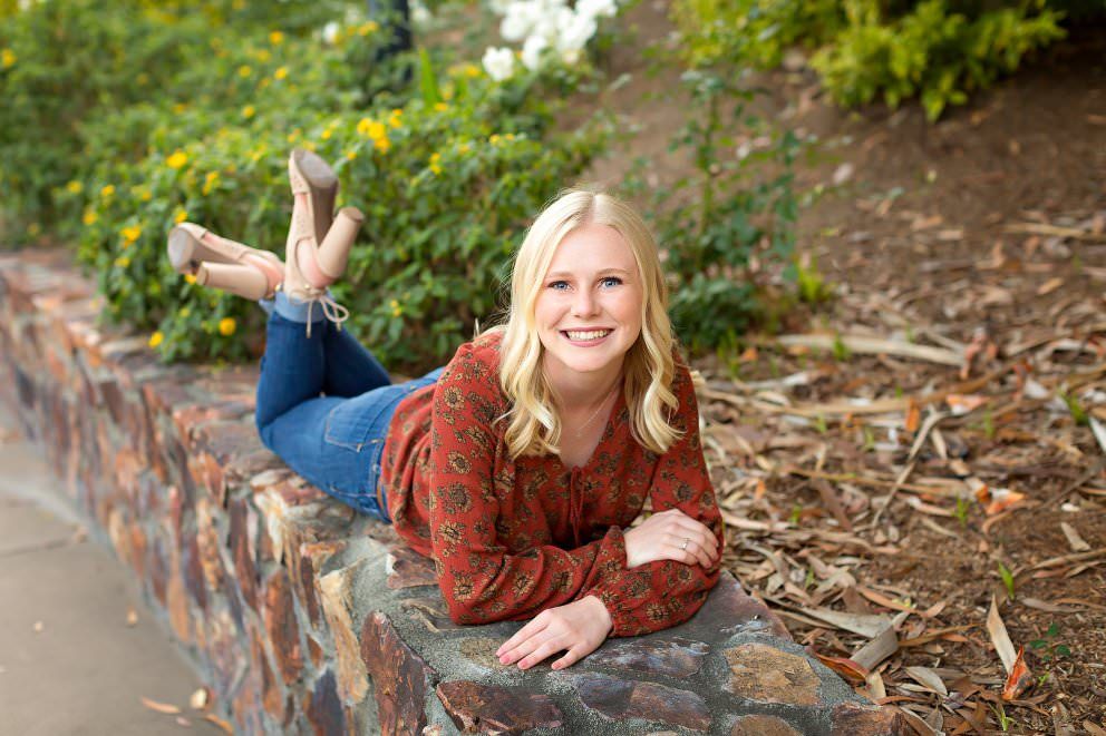 Smiling blonde teenage girl lying on a stone wall outdoors surrounded by lush greenery for senior portrait.