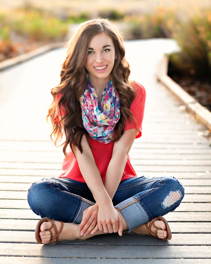 Young woman with long brown hair sits cross-legged on a wooden boardwalk, wearing a red top, colorful scarf, and ripped jeans.