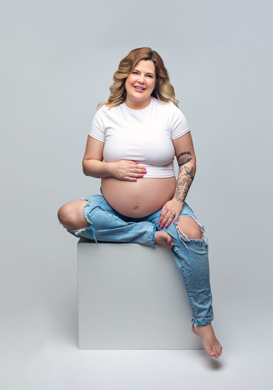 Pregnant woman in white crop top and ripped jeans sitting on white cube, showing baby bump with tattoo on arm.