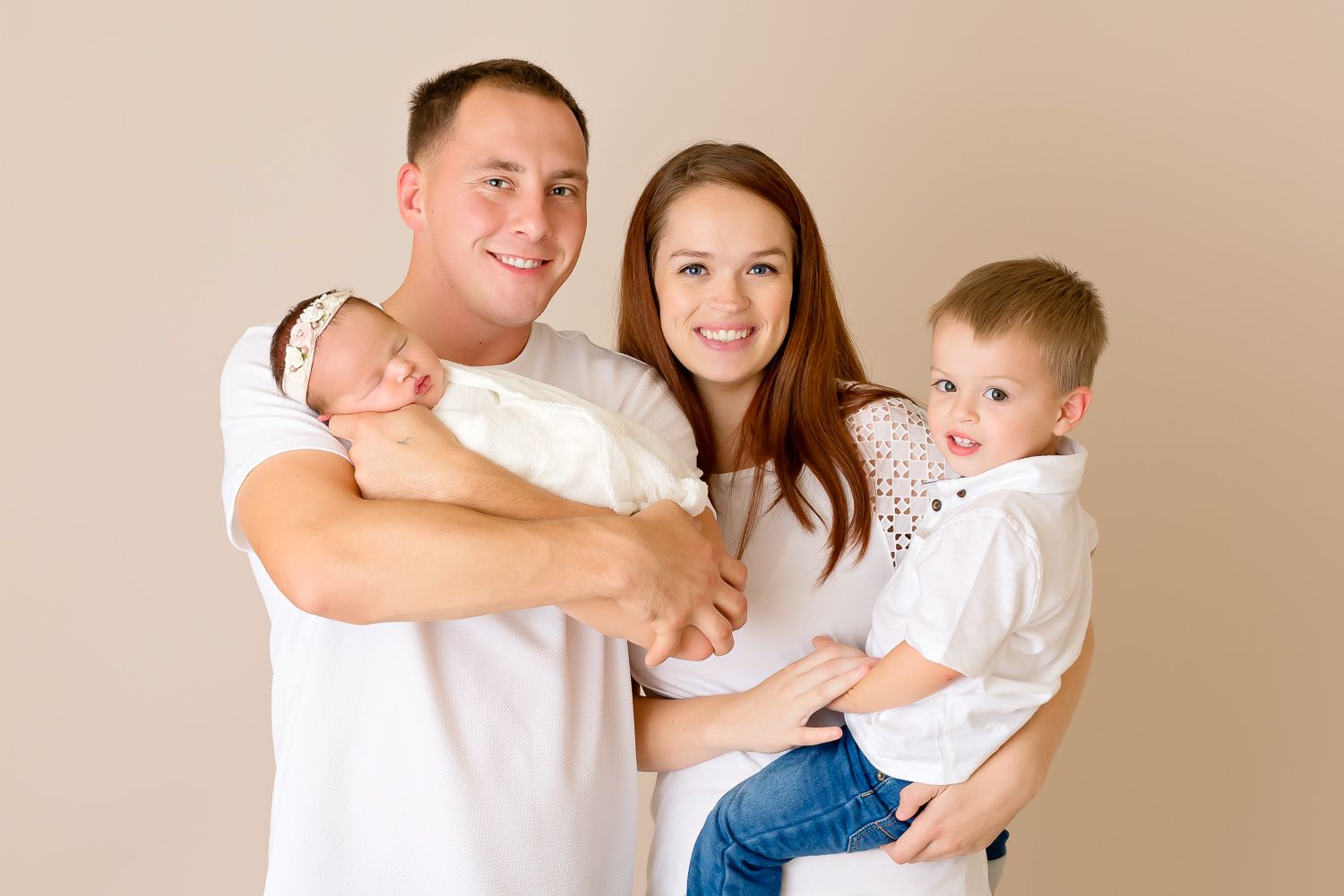 Happy family of four with newborn baby and toddler son posing together in white outfits.