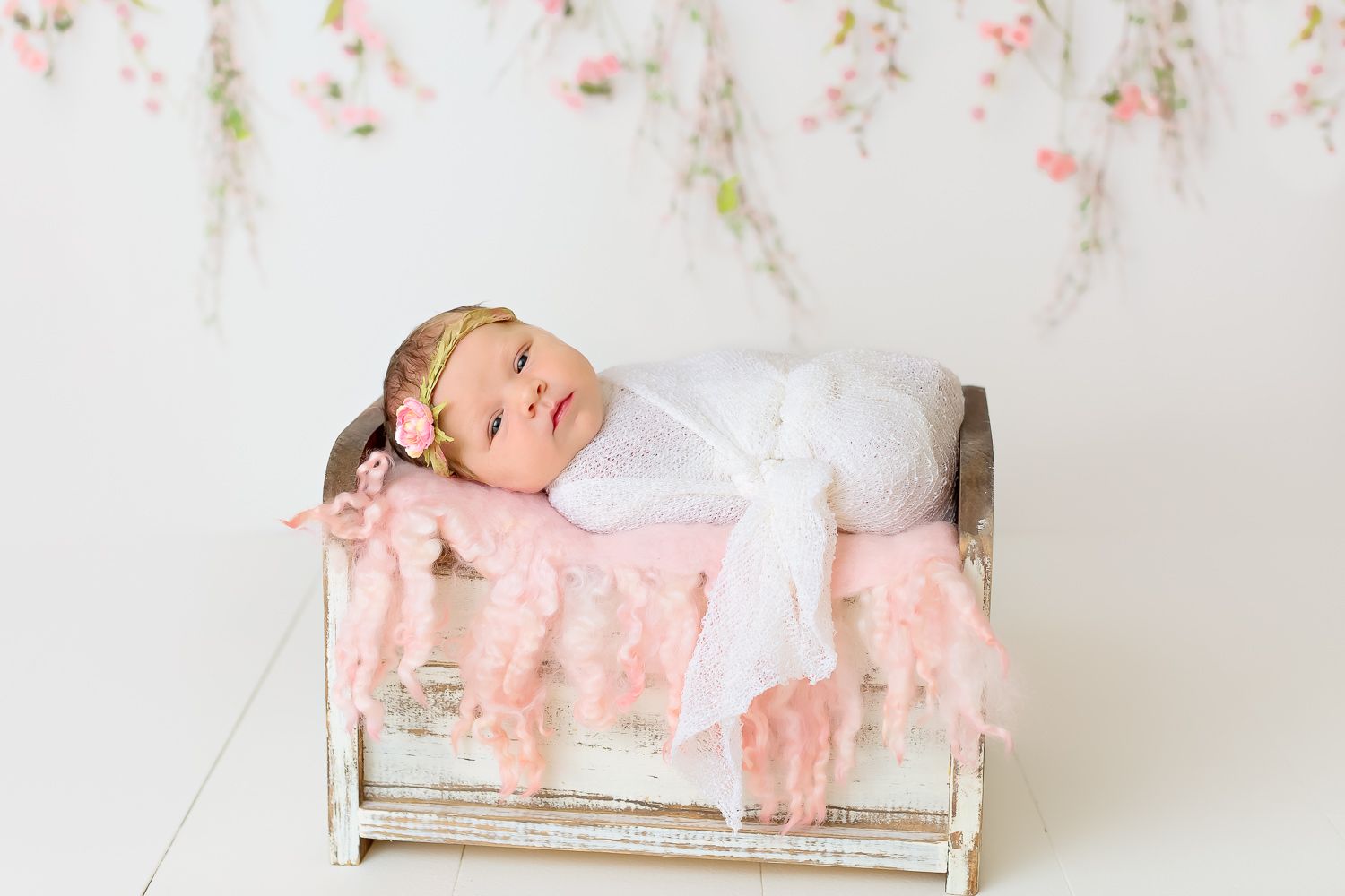 Peaceful newborn baby sleeping in a rustic wooden bed with soft pink feather blanket and floral backdrop.