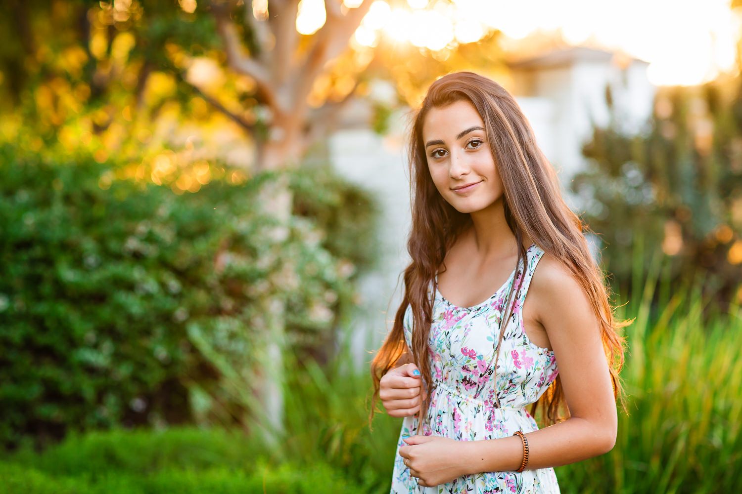 Young teenage girl with long brown hair wearing a floral dress, smiling outdoors in a sunny green garden.