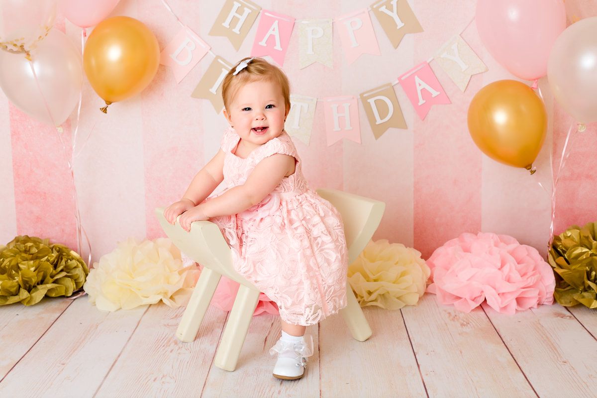 Happy baby girl in pink dress sitting on white chair with Happy Birthday banner and balloons for first birthday.