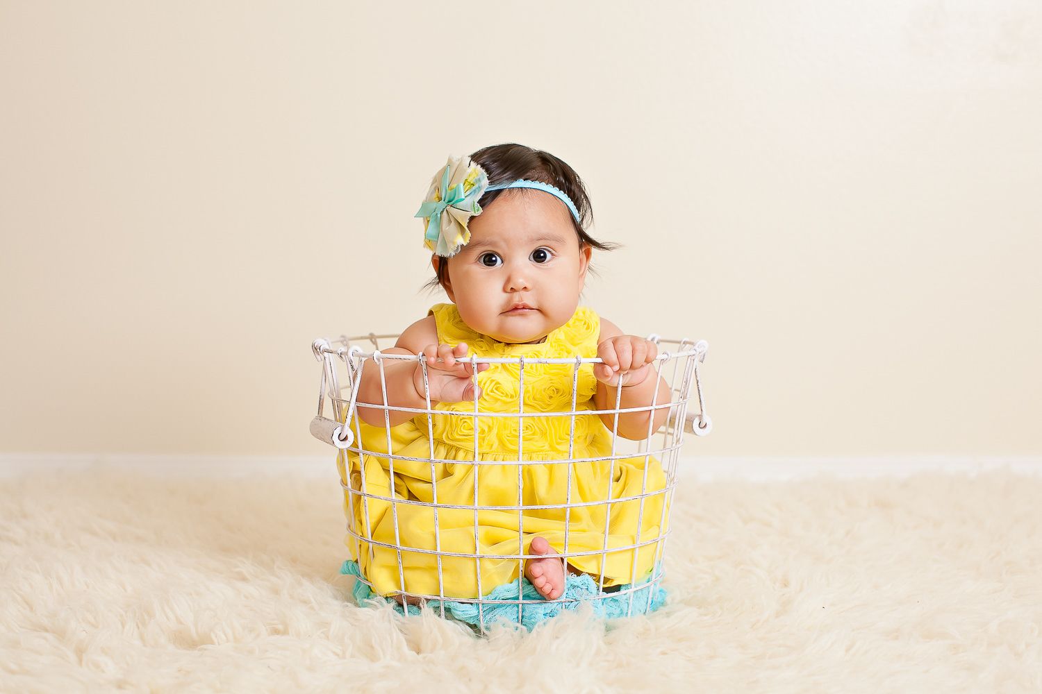 Cute baby girl in yellow dress and teal headband sitting in a wire basket.