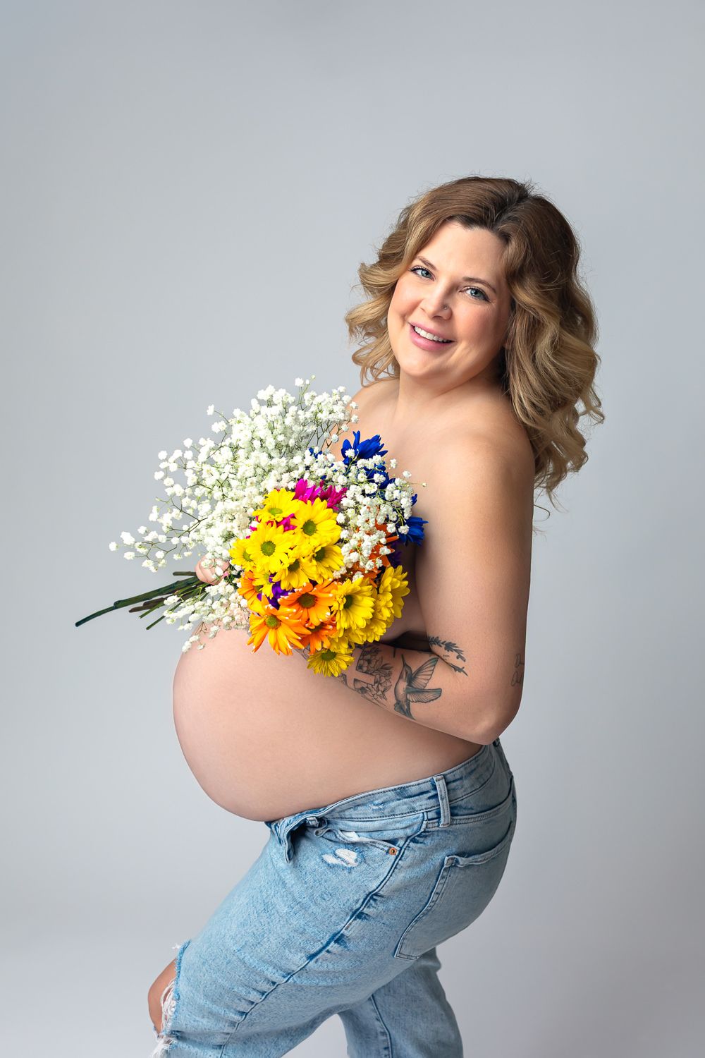 Smiling pregnant woman in jeans holding colorful flower bouquet, showing baby bump with tattoo in studio.