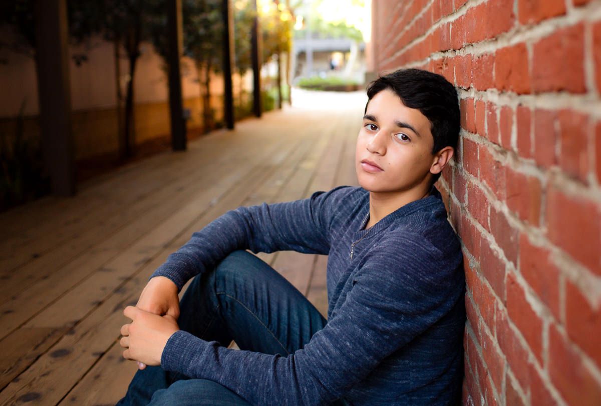 Teen boy in blue sweater sitting against red brick wall on wooden walkway, looking thoughtful.