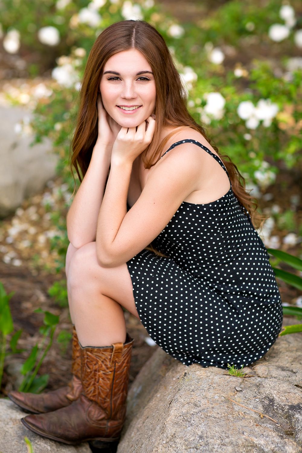 Young woman in black polka dot dress and cowboy boots sitting on rocks, smiling, with white flowers in background.