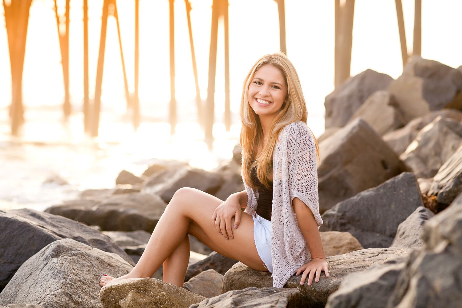 Young blonde woman smiling on rocky beach near pier at golden sunset, wearing white lace top.