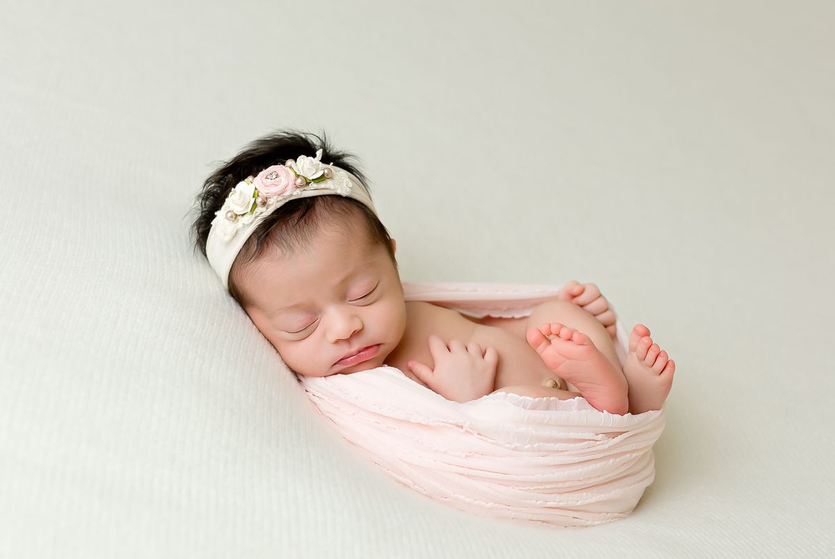 Newborn baby girl wrapped in pink swaddle with floral headband, sleeping peacefully on soft green background.