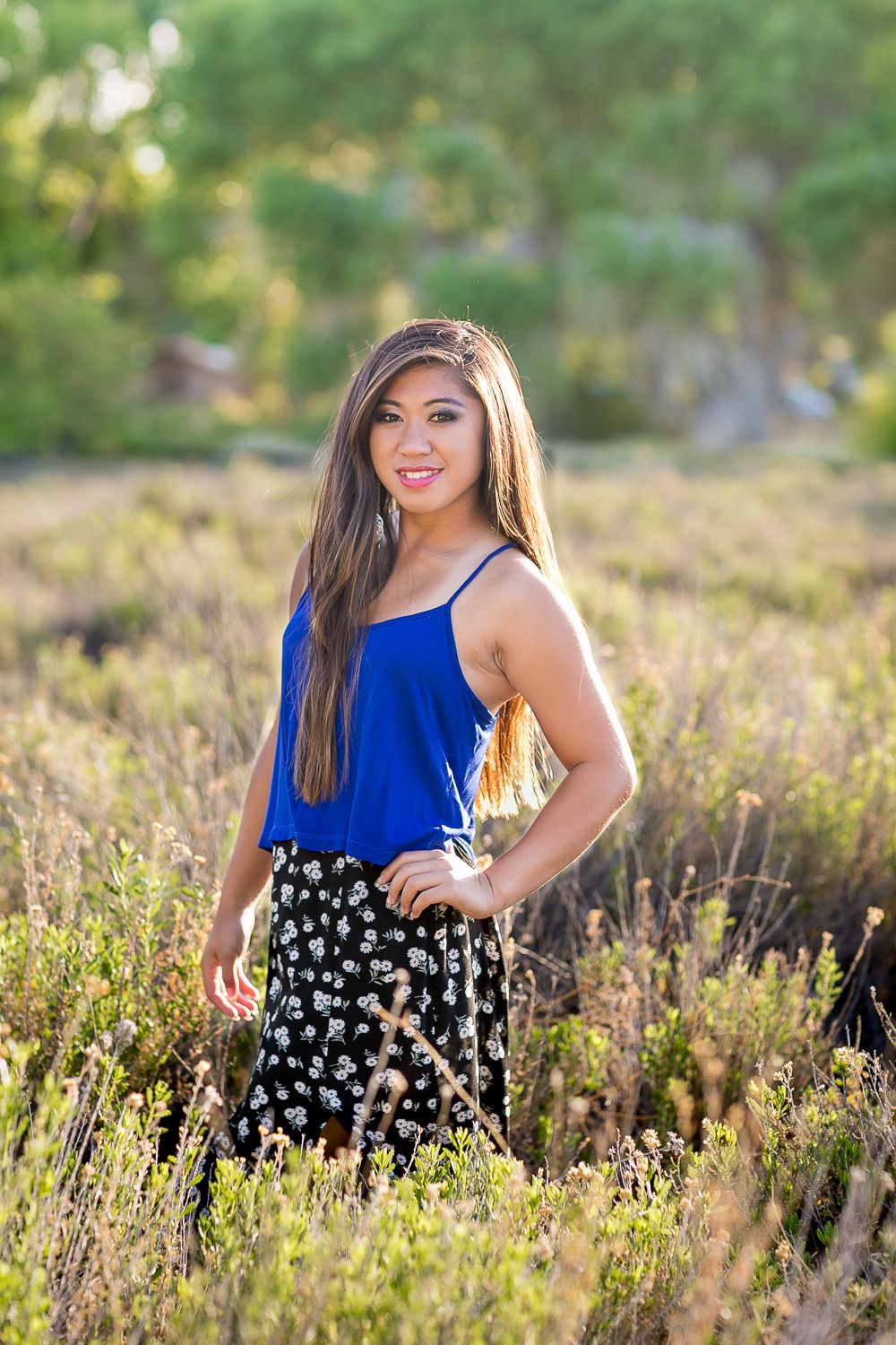 Young woman in blue top and floral skirt smiling outdoors in a sunlit field with green trees behind her.