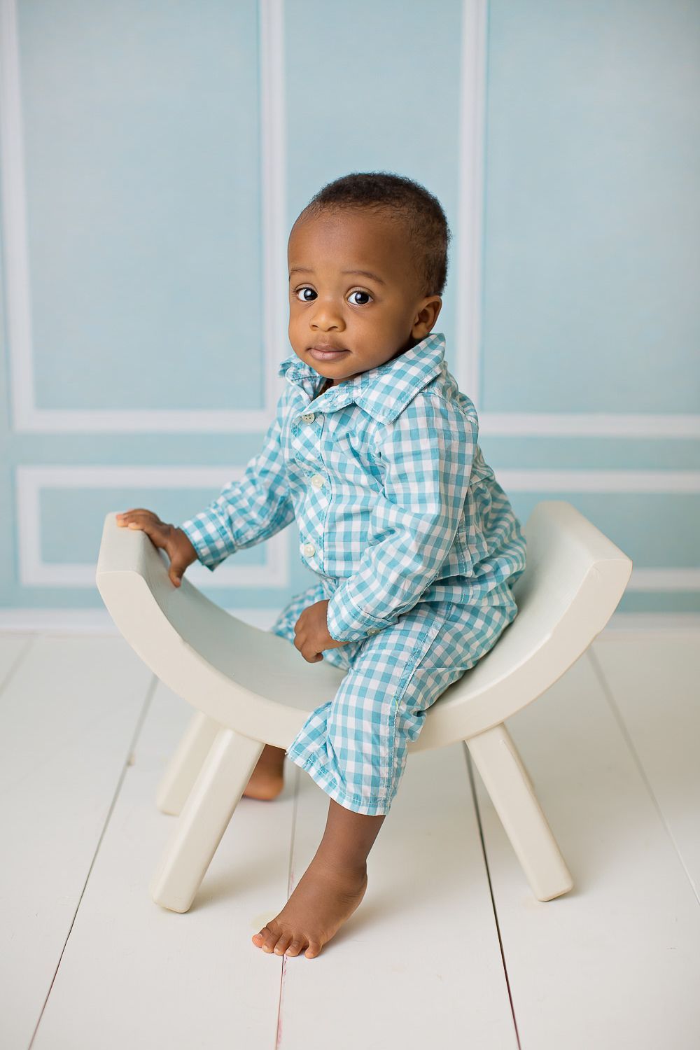 African American baby boy in teal plaid outfit sitting on white chair against light blue studio background.