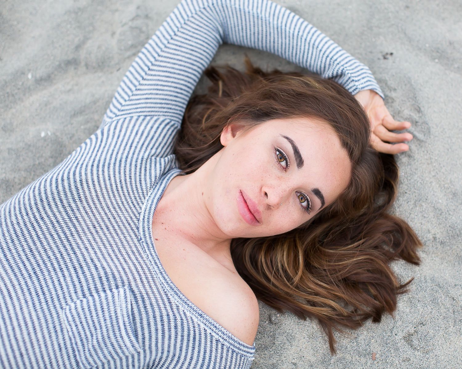 Young woman with brown wavy hair lying on sandy beach, wearing striped top, looking up at camera.