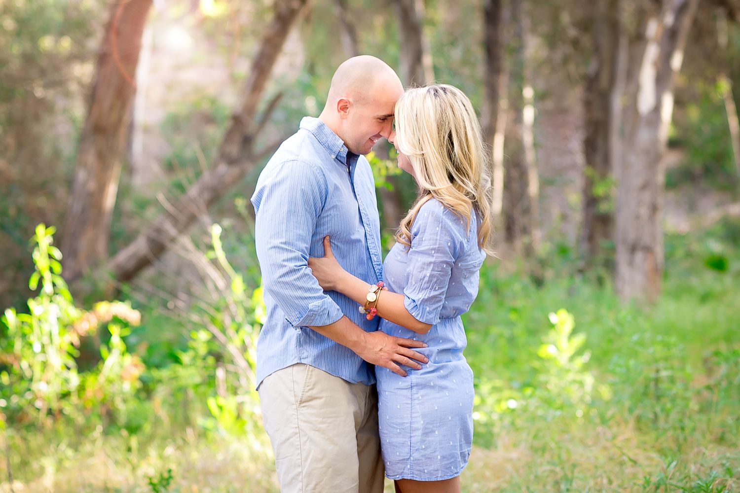 A couple embraces and leans in for a kiss in a lush green forest setting, both wearing matching blue outfits.