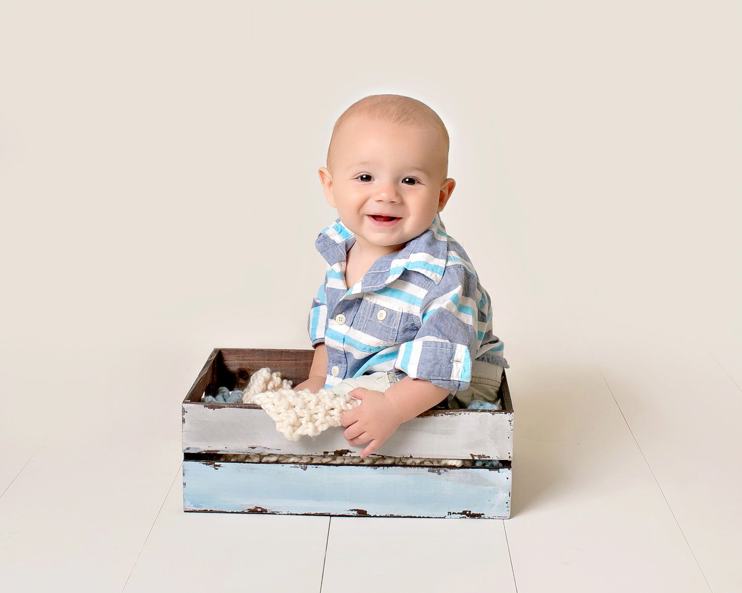 Smiling baby boy in plaid shirt sitting in a rustic blue wooden crate on a white floor.