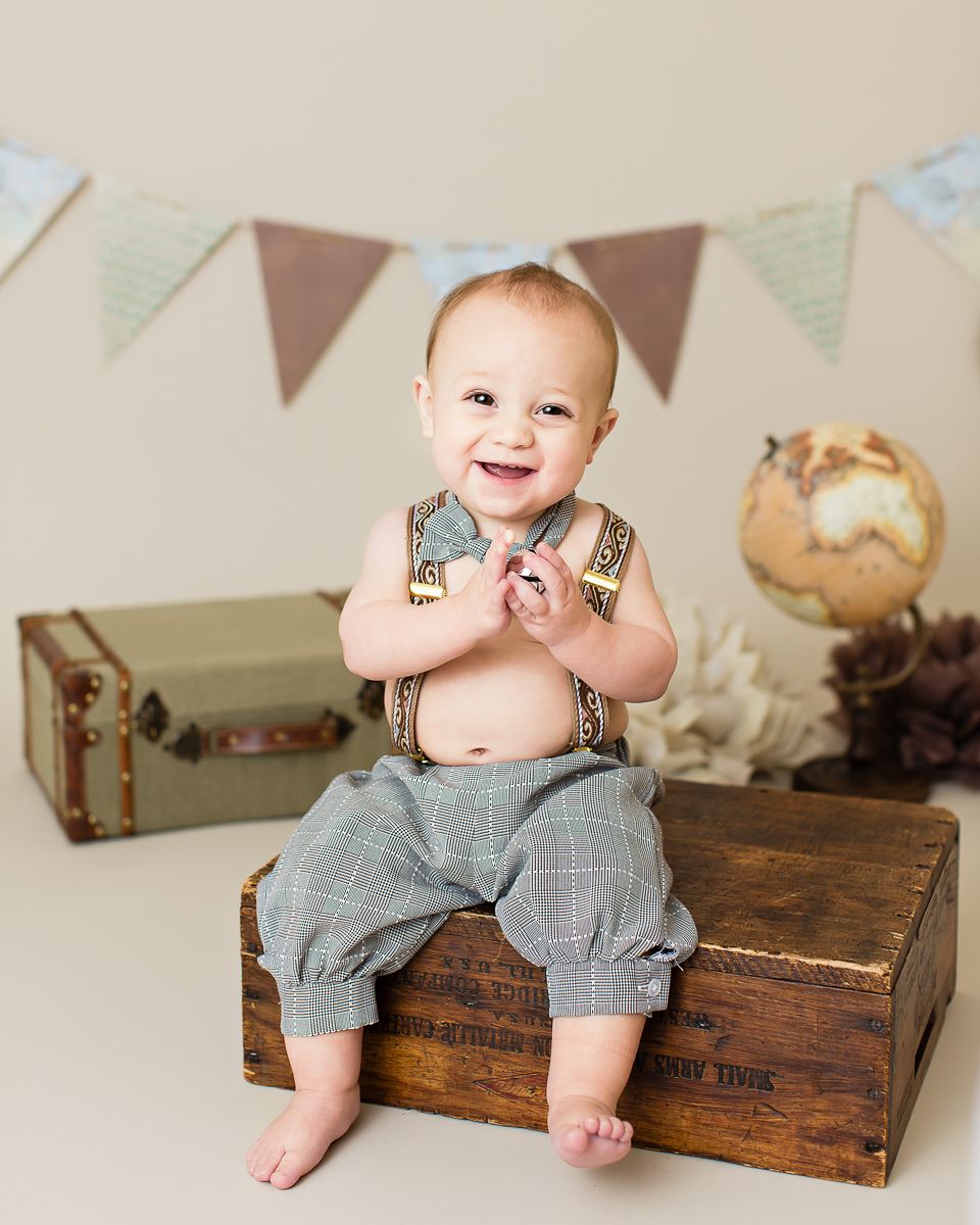 Smiling baby boy sitting on wooden crate wearing suspenders and plaid pants in vintage travel themed photo shoot.