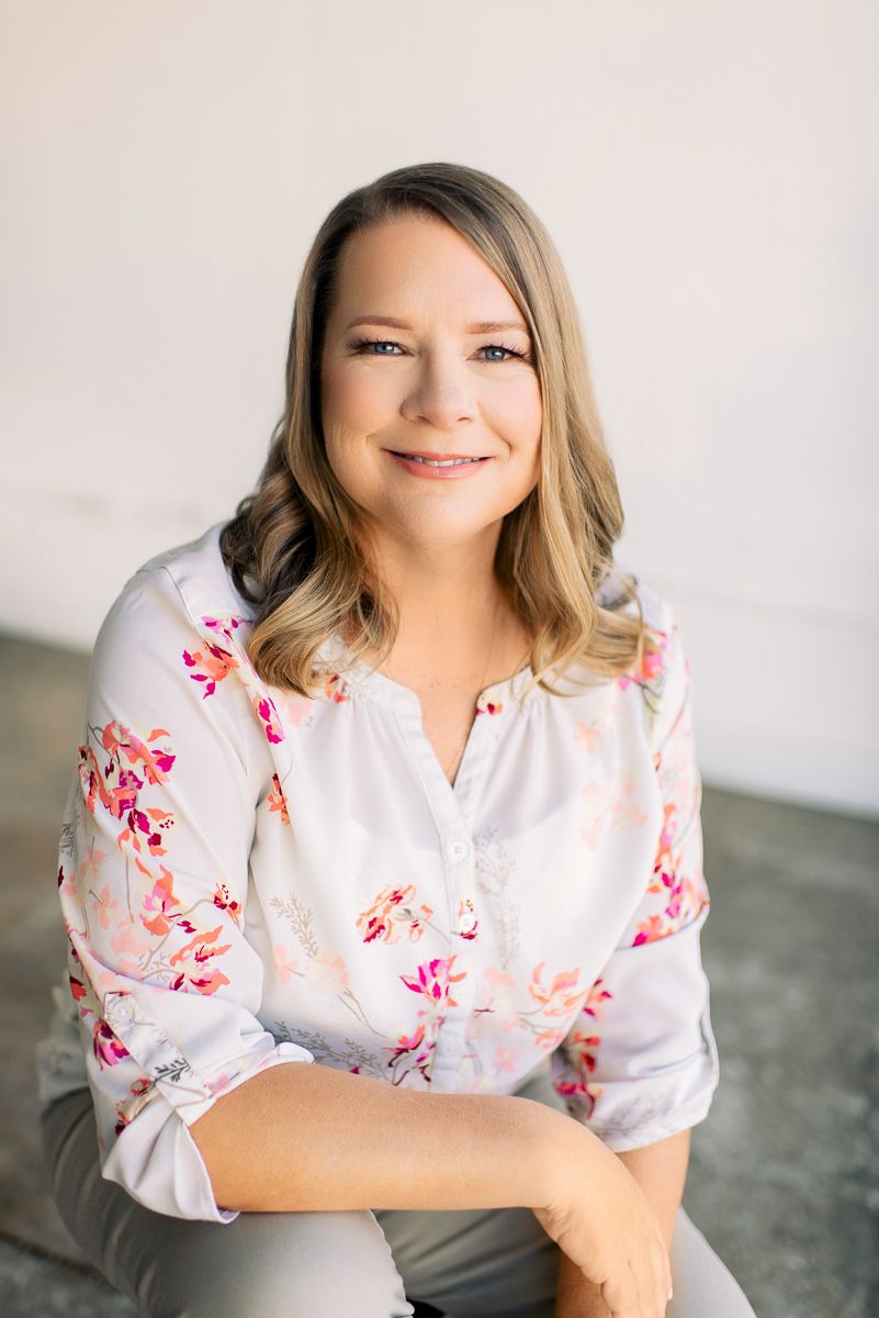 Smiling woman in floral blouse seated casually in bright studio portrait photography setting.