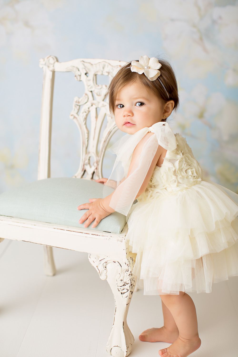 Baby girl in ivory tulle dress standing beside a vintage white chair.